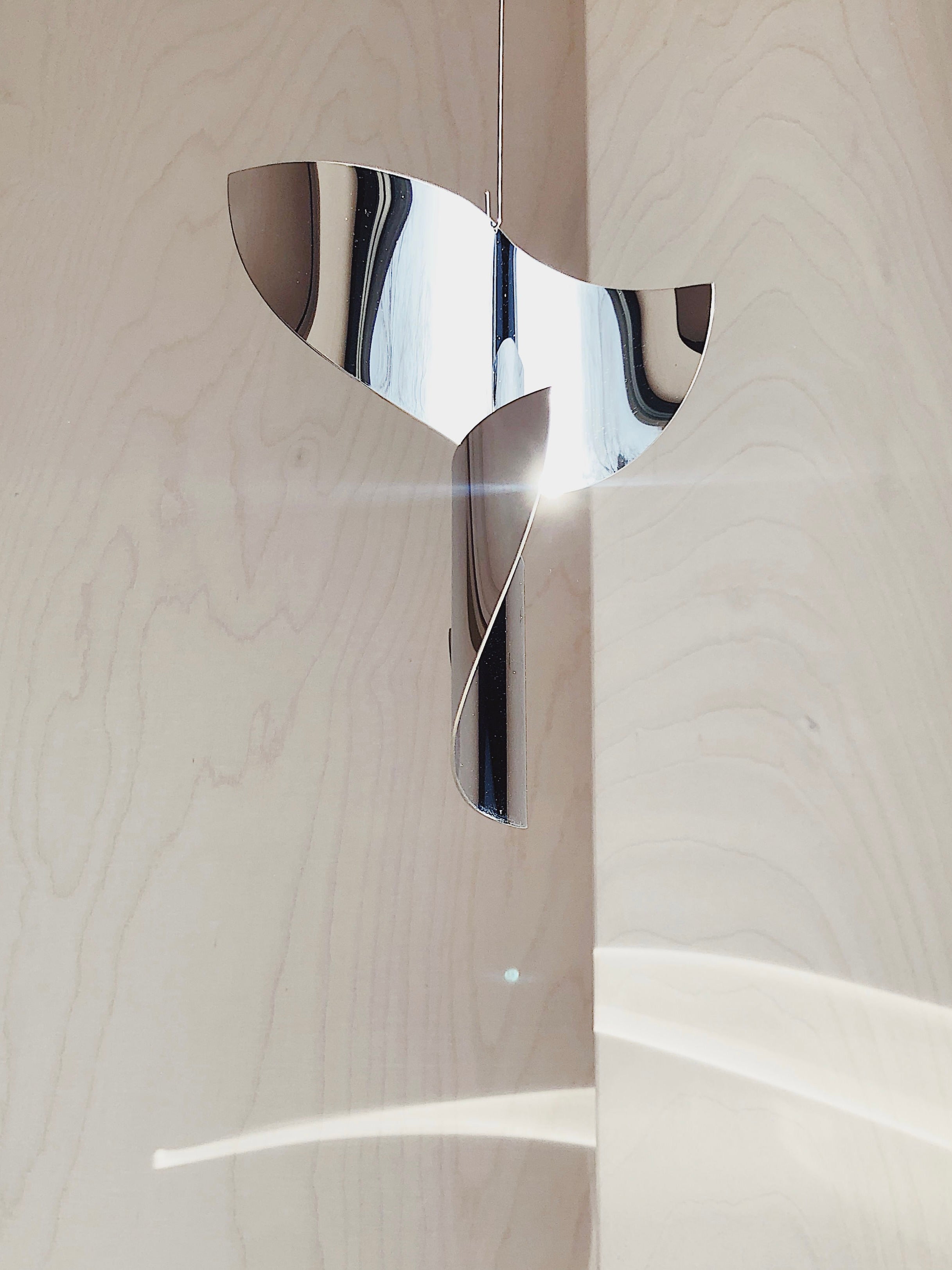 The Sun Catcher by Studio Fie Paarup is a handmade metallic abstract sculpture from Denmark. Its polished steel, twisted forms and modern minimal design reflect light and cast unique curved shadows against a pale wooden wall.