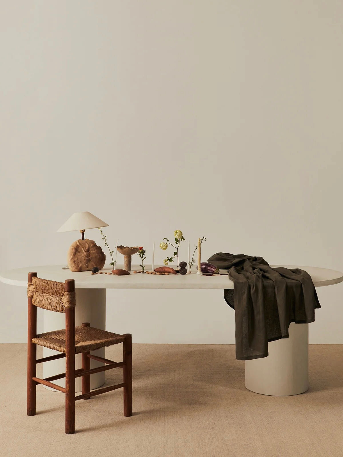 A minimalist dining area displays contemporary furniture with MarlotBaus's handcrafted wooden chair and an oval Reye Dining Table. A dark cloth covers the microcement table, adorned with a lamp, rocks, and small plants, set against a light wall and carpeted floor.