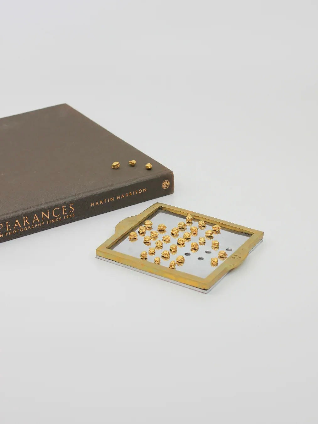 A brown book titled "Appearances - Photography Since 1945" by Martin Harrison lies flat on a white surface. Beside it is the English Solitaire, a rectangular decorative object crafted from brass and aluminum, with small gold accents reminiscent of David Marshall's style, available at Boga Avante Shop.