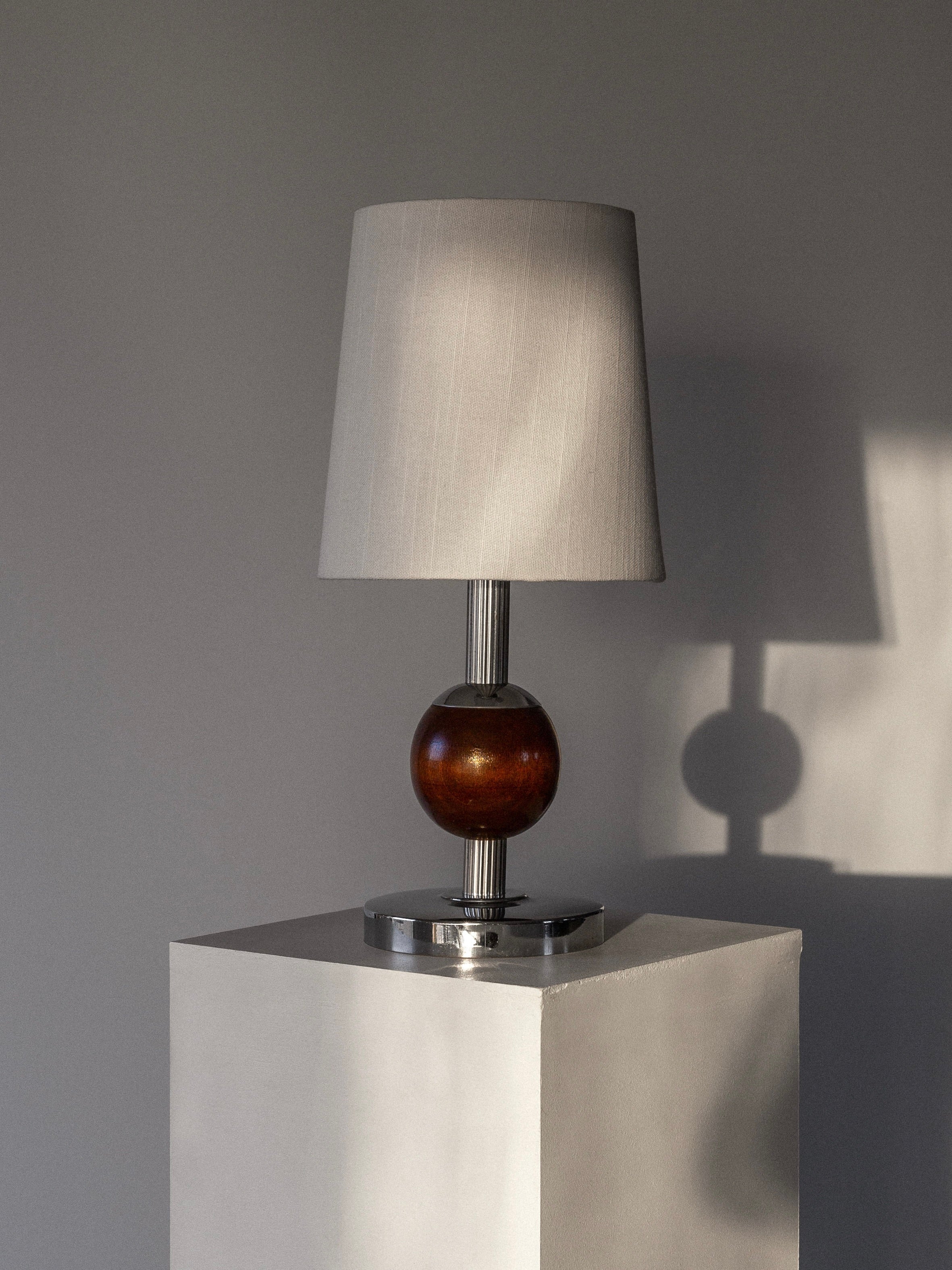 A Spigel Wood & Chrome Table Lamp from the 1930s, featuring a chrome and wood design with a beige shade and round wooden base, is positioned on a white pedestal against a gray wall. Sunlight casts its shadow onto the wall, evoking the style of 1930s Sweden.