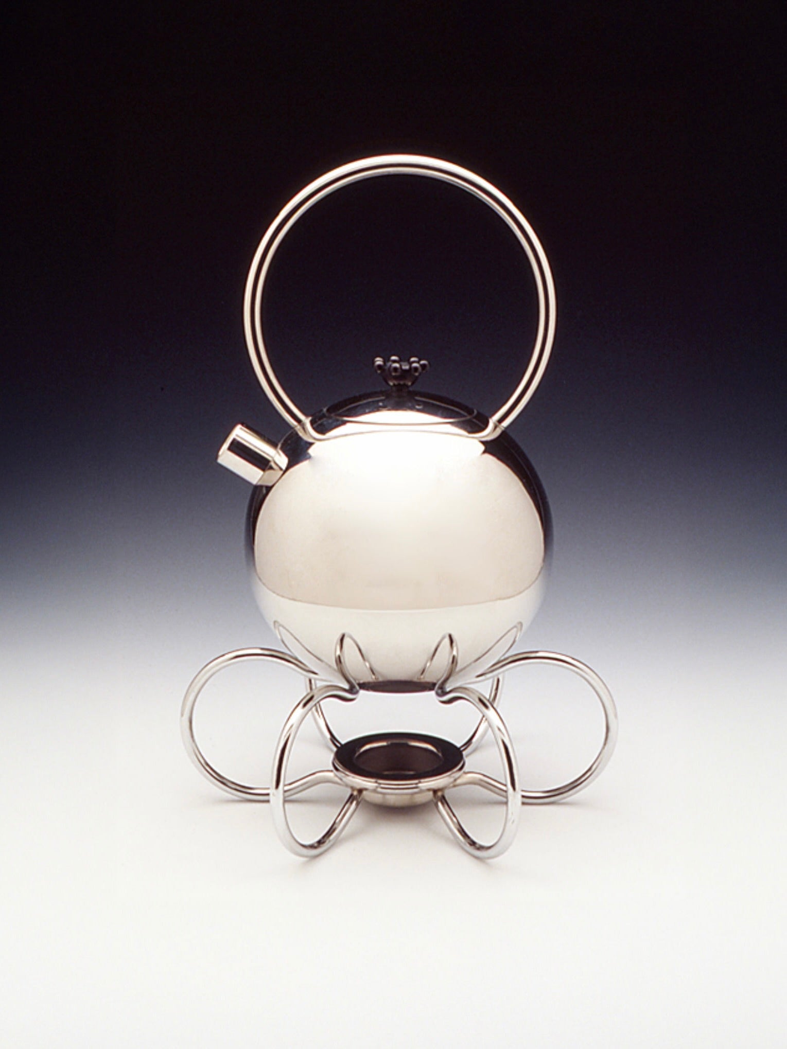 A modern Chrome teapot designed by Matteo Thun for WMF, featured by Dodo Vintage, boasts a spherical body and short spout. It rests on a stand crafted from coiled chrome Argan wires, forming a stylish and minimalist design. The large circular handle enhances its unique and contemporary aesthetic.