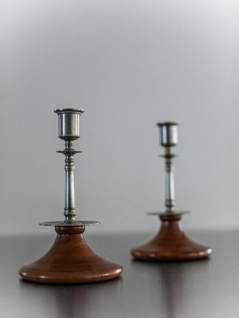 Against a plain backdrop, Out For Lunch's Steel & Wood Candleholders add rustic charm, featuring wooden bases and metallic holders. The closer candlestick is in focus on the dark surface, while the other is slightly blurred.