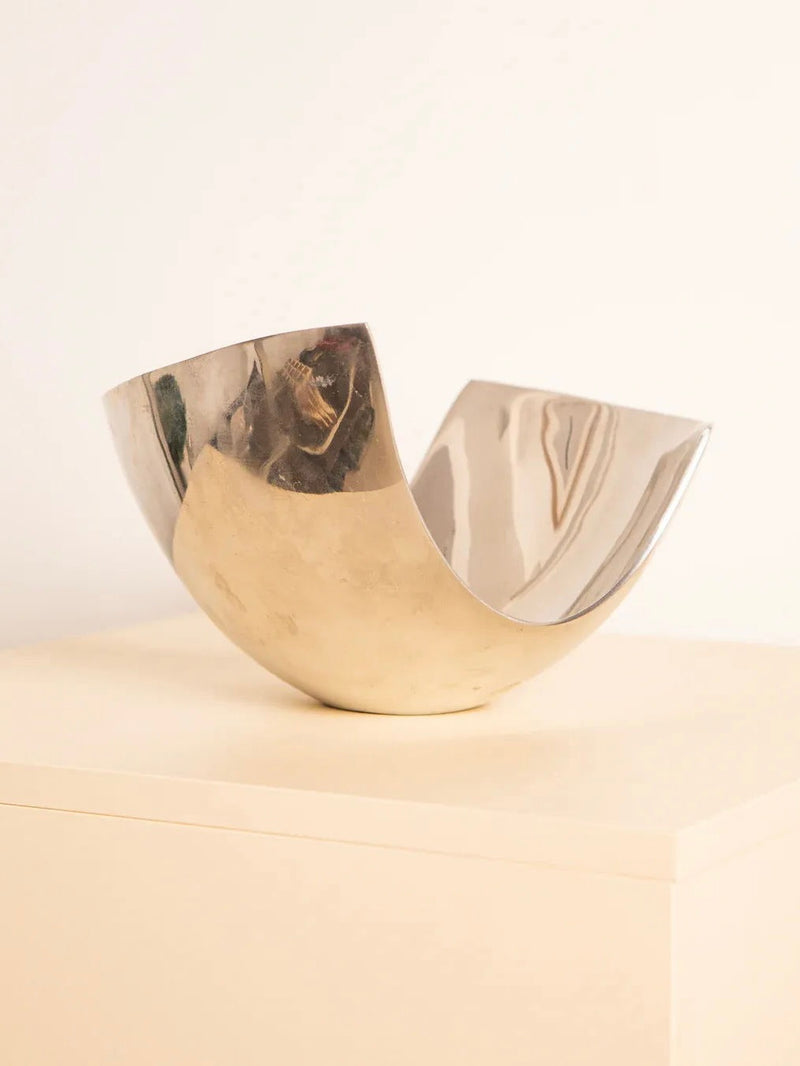 A "U"-Shaped Fruit Basket 80's, crafted by Treaptyque, features a gleaming metallic finish with a reflective surface. It rests elegantly on a light-toned rectangular stand against a simple backdrop, showcasing an asymmetrical, abstract form inspired by retro design aesthetics.