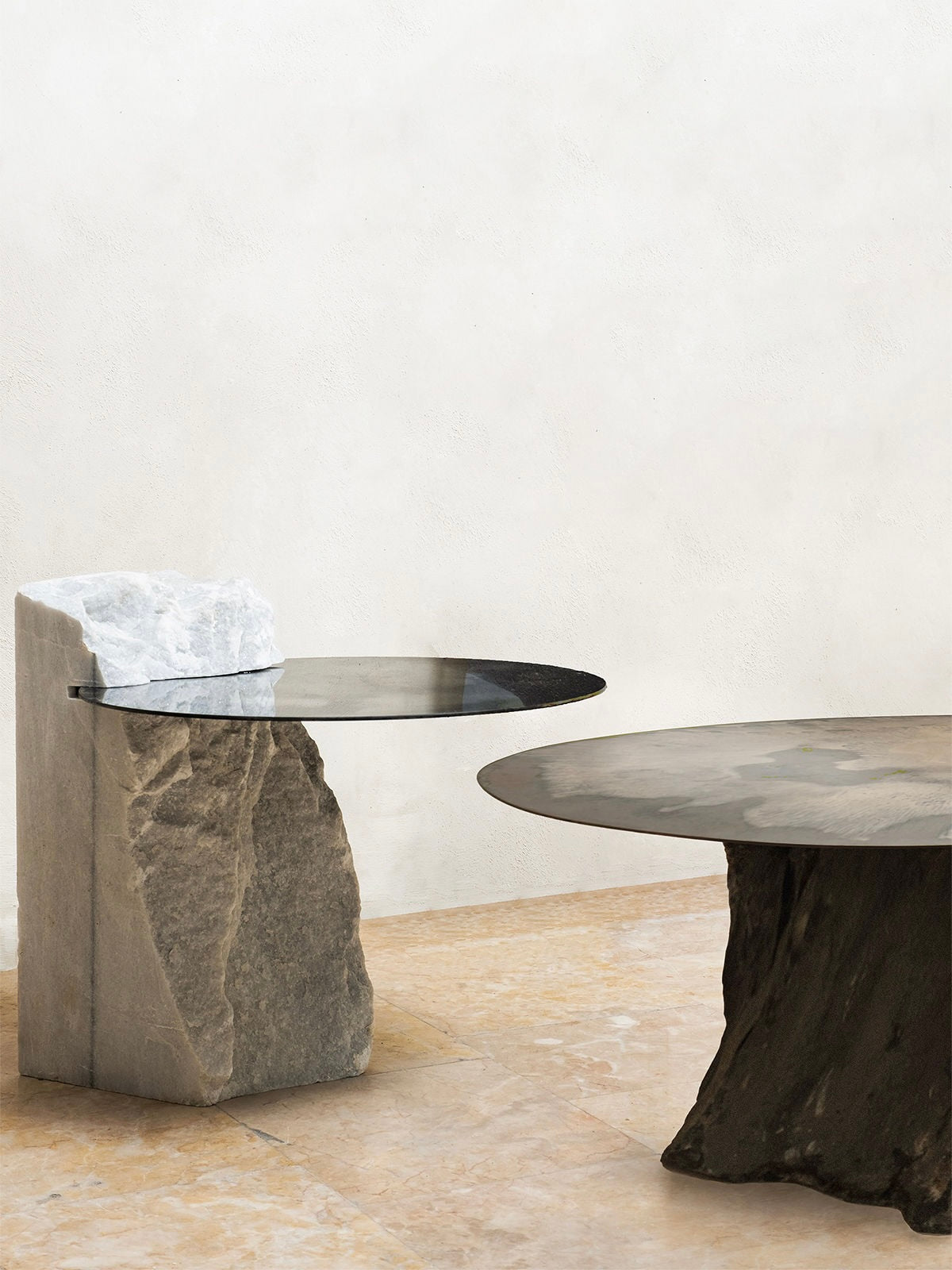 Against a plain wall, two HOMA Cloud Mirror Side Tables stand: one has a tall, rough stone base topped by a small round glass top, the other is lower with a wide circular glass surface on an elegant marble-textured foundation.