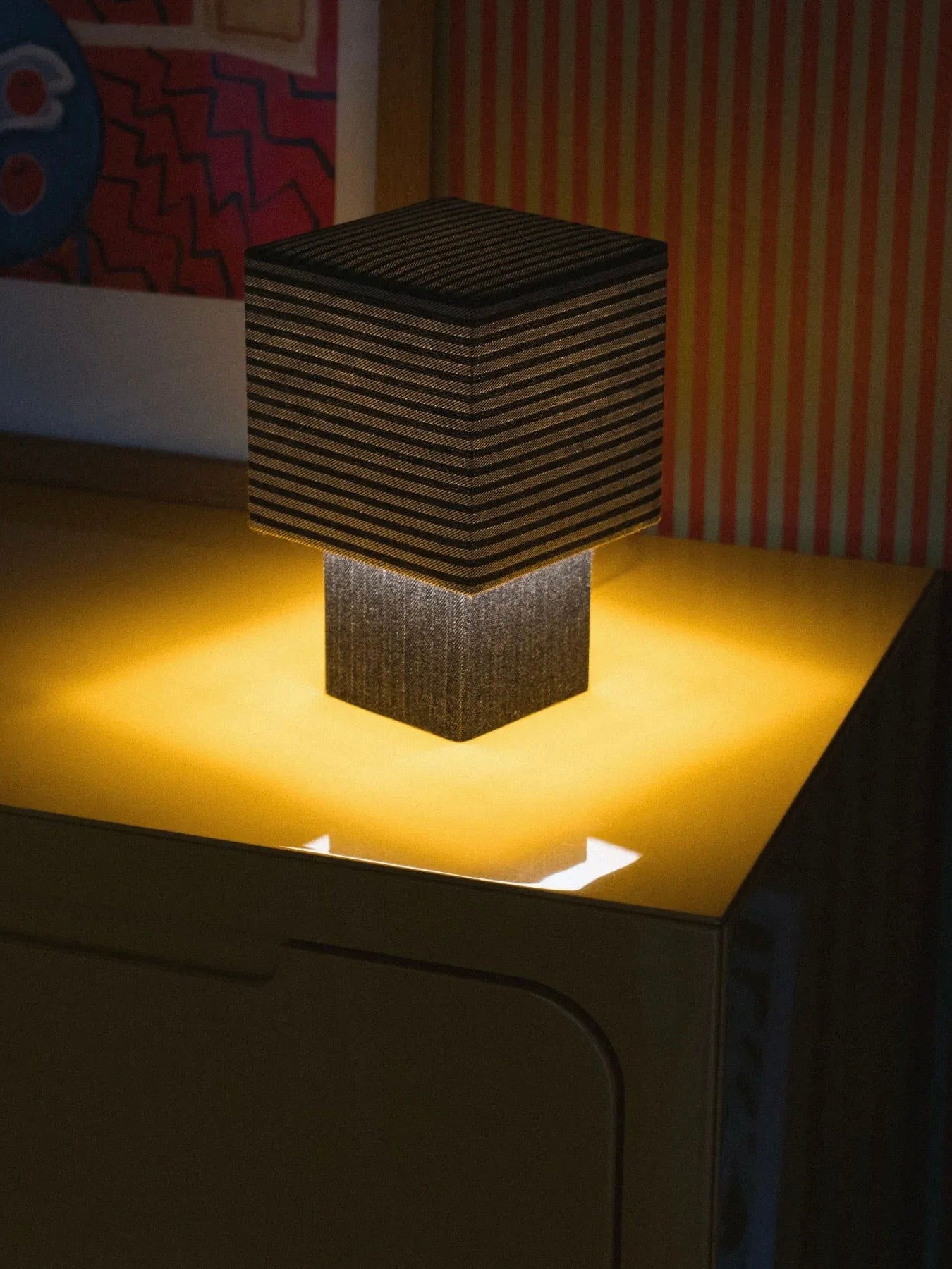 The "La Petite Boîte" Striped Denim Table Lamp by mattina moderna, a modern square-shaped lamp crafted from deadstock fabrics, emits a warm light with its horizontal stripes and rests on a yellow table. In the background, abstract artwork displays red zigzag patterns and vertical pink stripes. This handcrafted piece embodies environmental consciousness in design.