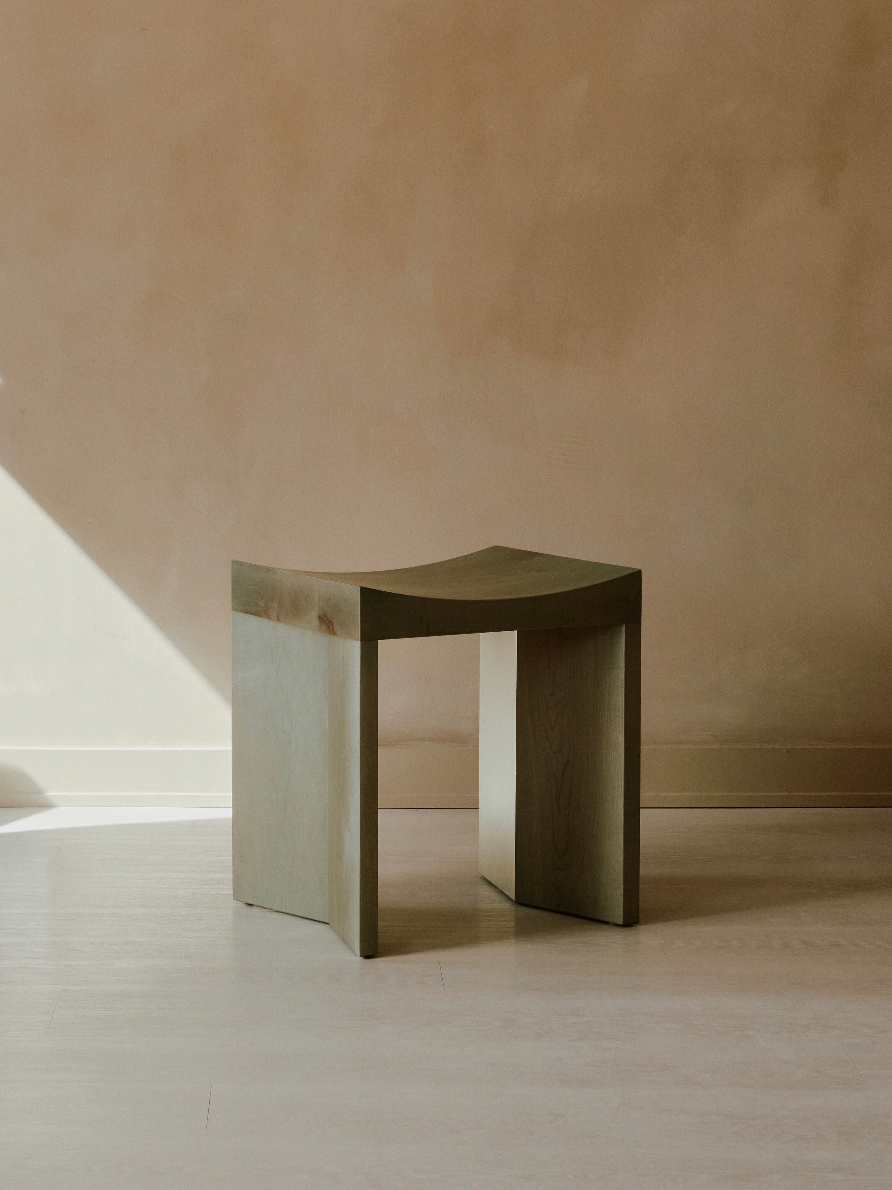 An Arc Stool by Juntos Projects, featuring a minimalist wooden design with a slightly curved seat and flat, rectangular legs, is placed in a room with soft, natural light. The background showcases a beige textured wall, creating a warm and serene atmosphere.