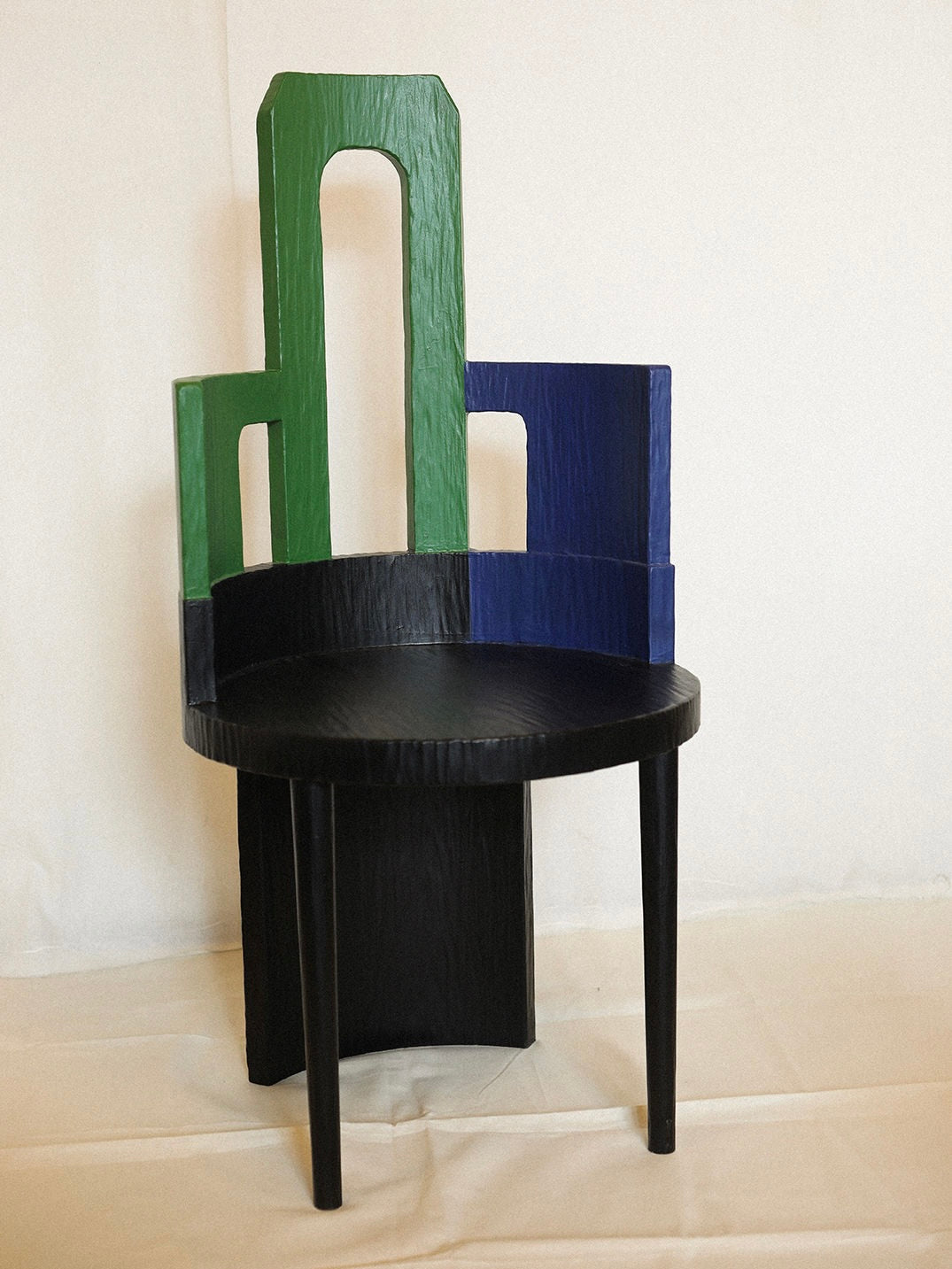 The Sculptural Chair III in Primary Colors by Rooms Studio features a black round seat and a wooden backrest with vertical geometric shapes in green and blue, crafted from reclaimed oak, and is displayed on a light-colored surface against a plain backdrop.