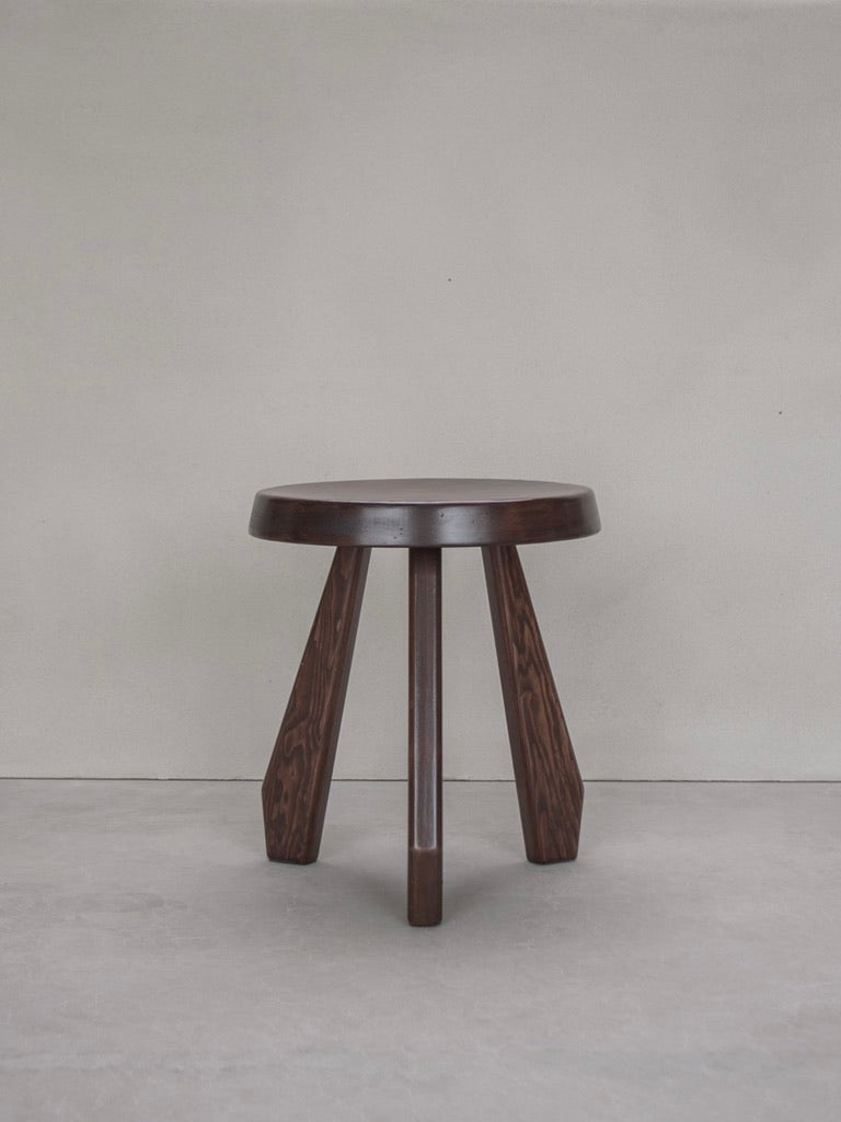 The Bicci de' Medici Charlotte Perriand - Sandoz Stool, featuring a round seat and three angled legs, stands out against a gray wall and floor. This mahogany piece has a dark polished finish that brings timeless elegance to any setting.