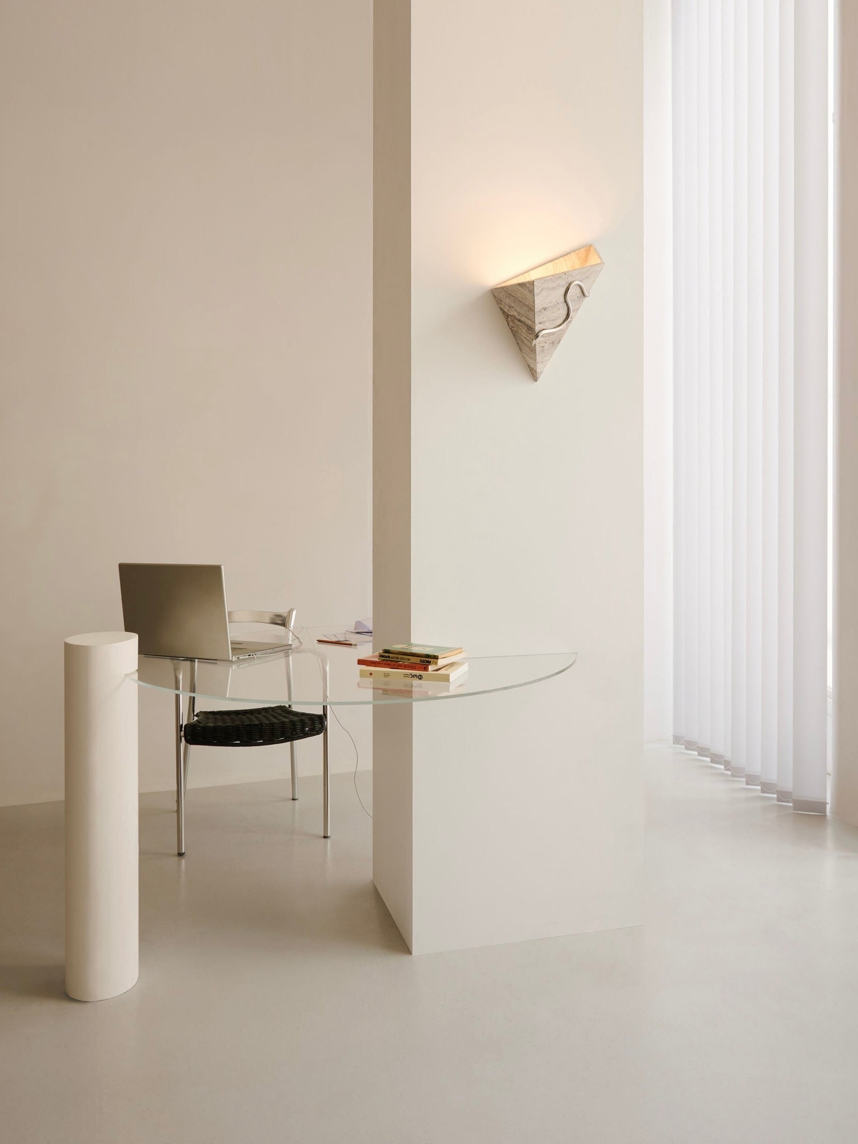 A minimalist office space showcases a sleek glass desk mounted to the wall, paired with a modern chair featuring a black seat and complemented by a silver laptop. A vertical cylindrical column enhances the foreground, while a geometric wall sconce and the Mariza Galani Therapeutic Wall Light 3 introduce an elegant touch with their indirect marble illumination.