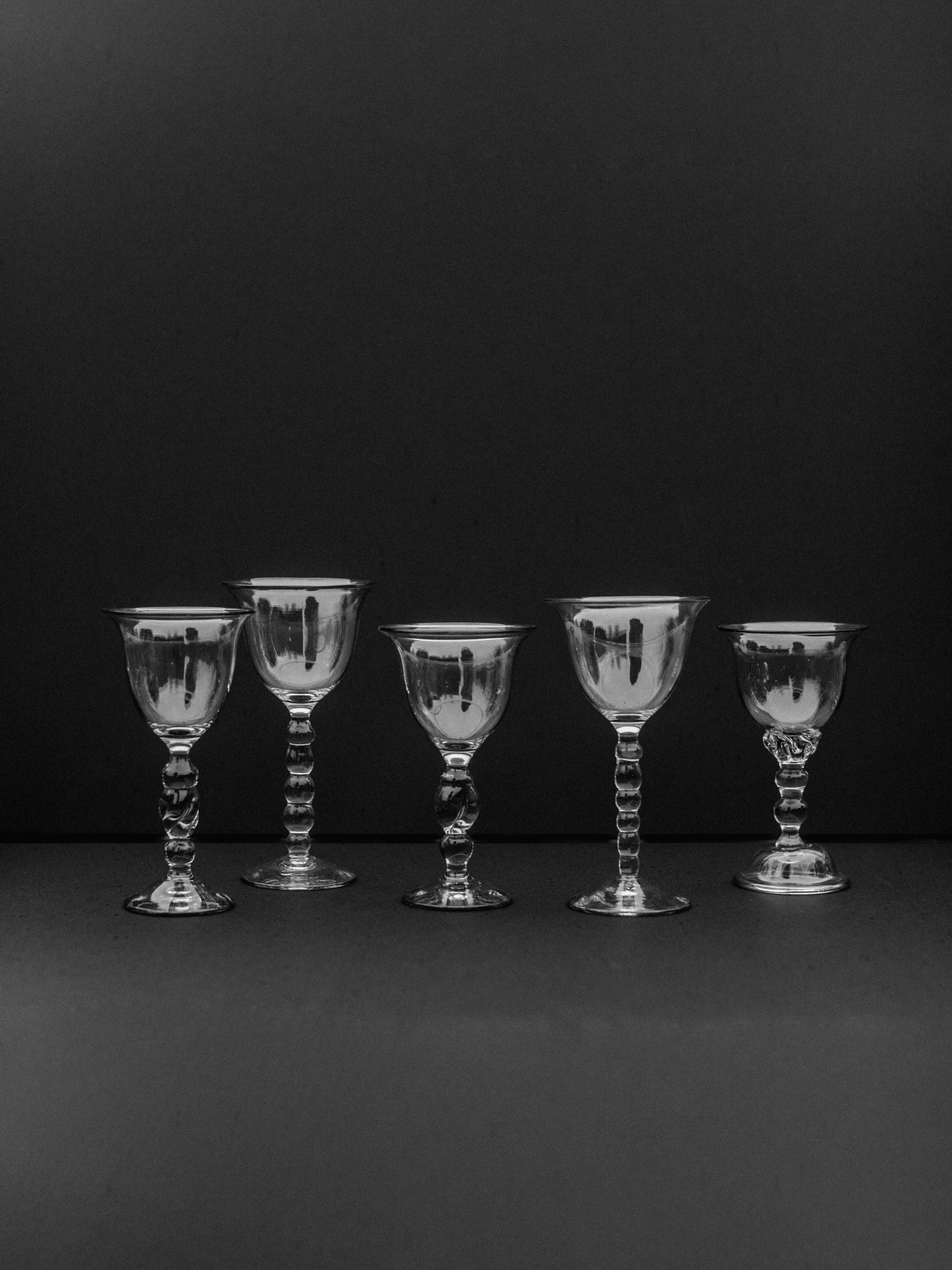 A set of five handmade art glasses by Out For Lunch features vintage-style goblets with ornate stems, arranged in a row against a dark background. The glasses showcase artisanal craftsmanship, with reflective surfaces creating subtle highlights and shadows.