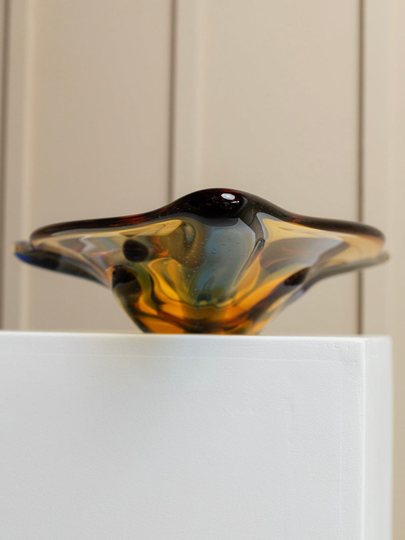 A close-up of the Vintage Glass Bowl by Karel Zemek from Bottega Jacobs reveals its glossy, multicolored surface, reminiscent of classic retro glassworks. The smooth curves display a harmonious blend of colors ranging from amber to dark brown with subtle hints of blue. The piece rests elegantly on a white pedestal against a light-colored background.