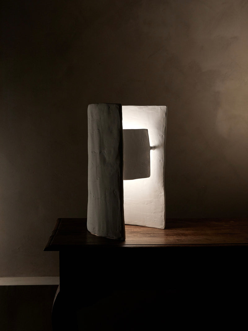 A Studio Tristan Kallas Crescent Table Lamp, sculptural in design with an open rectangular frame and cylindrical section, sits on a wooden table. It casts warm, indirect light and shadows on the wall, creating a minimalist setting with soft ambient lighting.