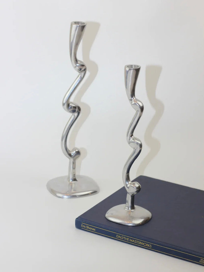 A pair of Boga Avante Shop aluminum candleholders, featuring a sculptural and wavy design, elegantly rests on the hardcover book "Dali: The Masterworks" by Eric Shanes. These pieces contribute a modern flair against the plain white background.