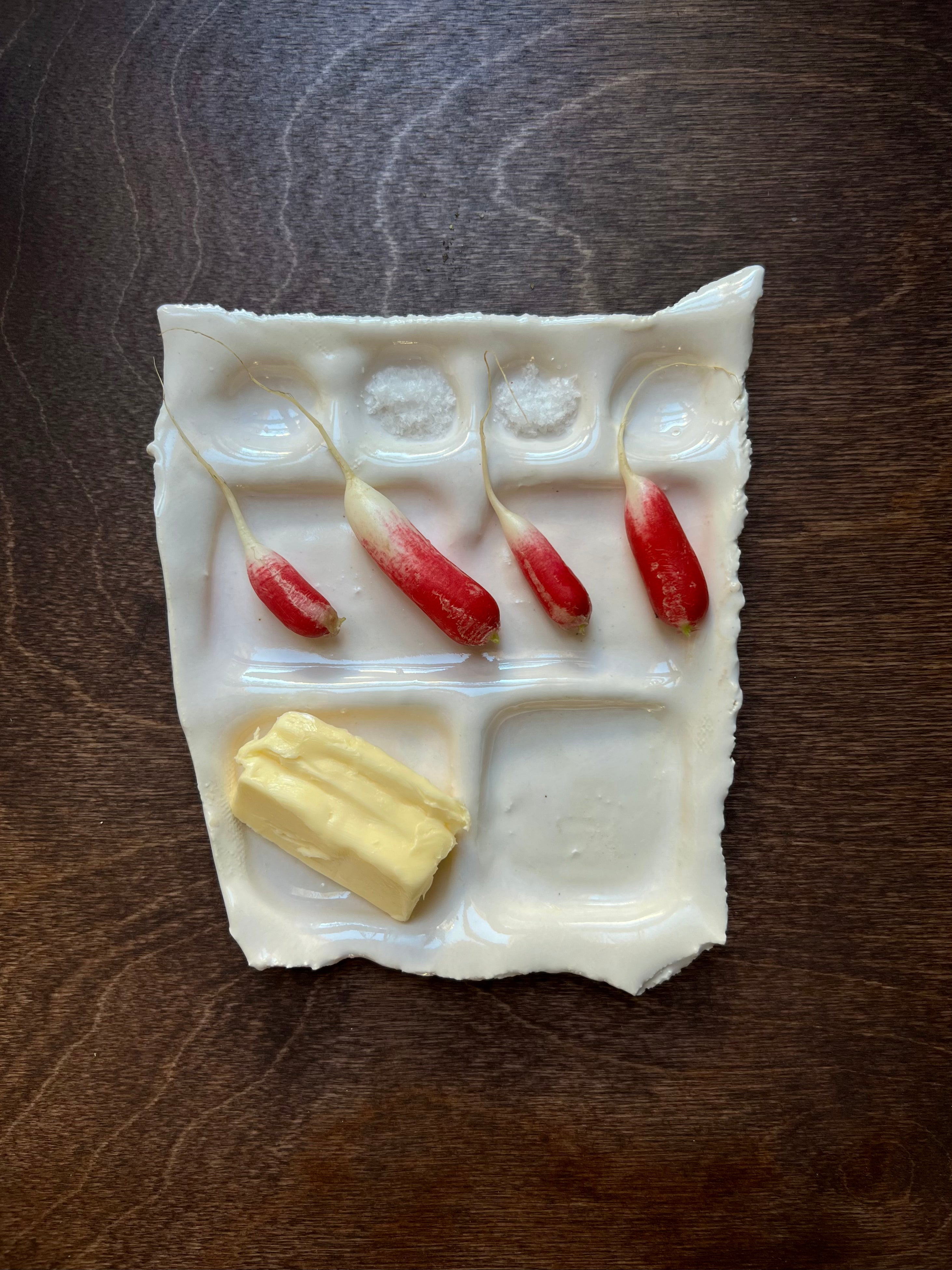 An image of a white clay Snack Plate by Somthings containing a stick of butter and three miniature red peppers. The Snack Plate, featuring a shiny white glazed finish, rests on a dark wooden surface.