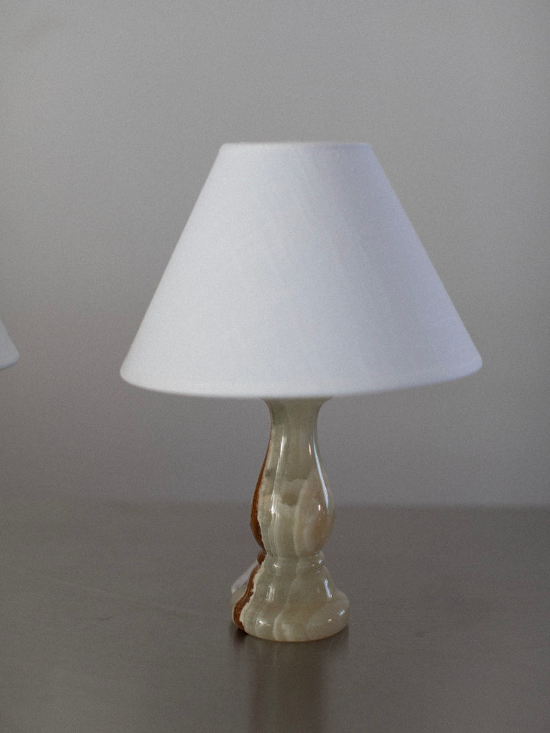 A small table lamp from the Out For Lunch Marble Table Lamps Duo collection, featuring a white conical lampshade and an onyx marble base, sits on a grey surface. The base showcases a smooth, curvy design with shades of beige and brown. The background is a plain, light-colored wall.