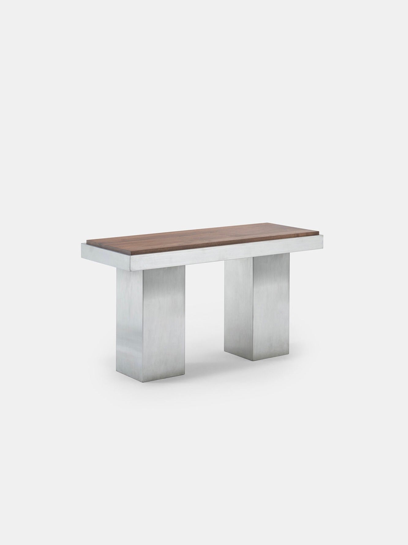 The Console Table 01 from Studio Vraco is a minimalist piece inspired by Bauhaus design, featuring a wooden top and two broad, rectangular legs made of brushed stainless steel, all presented against a plain white backdrop.