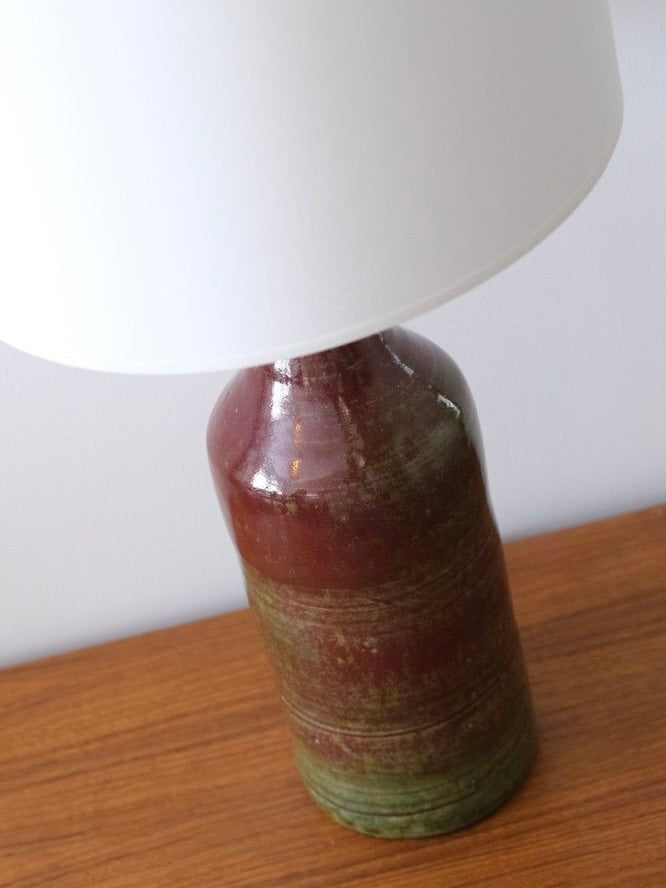The Ceramic Lamp by Fongombault from LA NOUVELLE GALERIE features a 70's style with a glazed brown and green base. It sits on wood, topped with a white cotton lampshade that enhances its dark red to earthy green tones, evoking the serene simplicity of Abbaye de Notre-Dame de Fongombault.