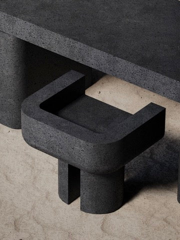 A minimalist Monolith M_001 Chair, dark gray concrete with a throne-like form, U-shaped backrest, and two sturdy legs, sits before a matching table. The light beige stone background contrasts strikingly with the furniture.