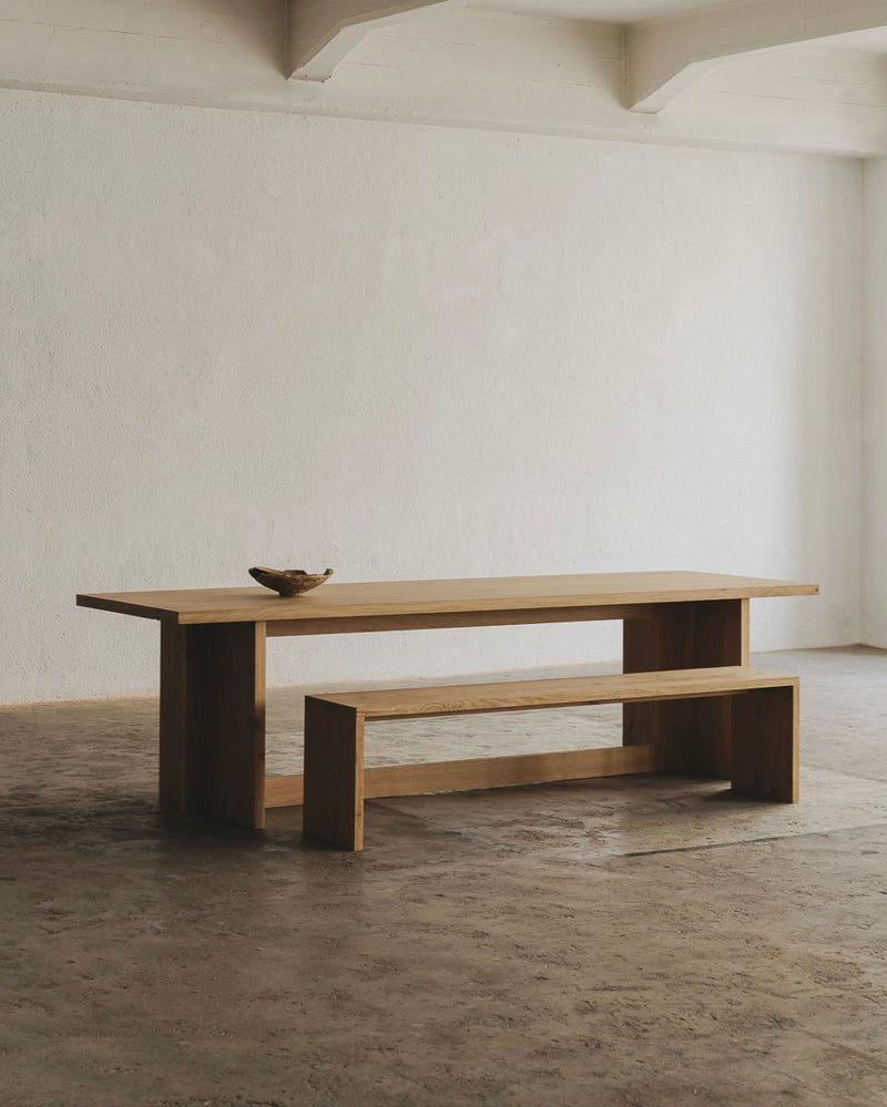 A minimalist, spacious room showcases the Paris Dining Table from ASERIES OF OBSTACLES, SL with a matching bench for social gatherings. A small decorative bowl accents the table against textured concrete floors and white walls, with an exposed beam. Natural light enhances French elegance.