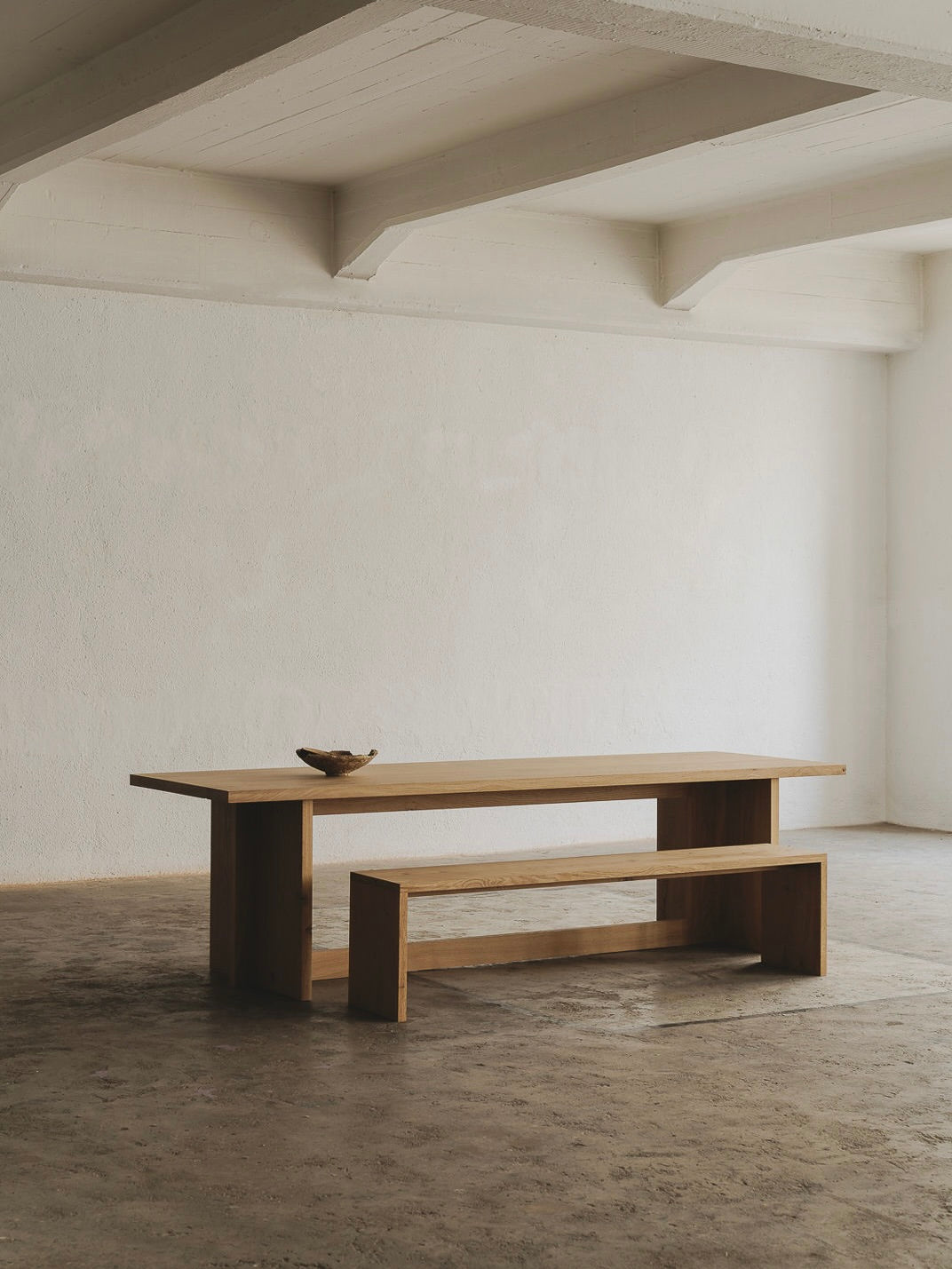 A minimalist, spacious room showcases the Paris Dining Table from ASERIES OF OBSTACLES, SL with a matching bench for social gatherings. A small decorative bowl accents the table against textured concrete floors and white walls, with an exposed beam. Natural light enhances French elegance.