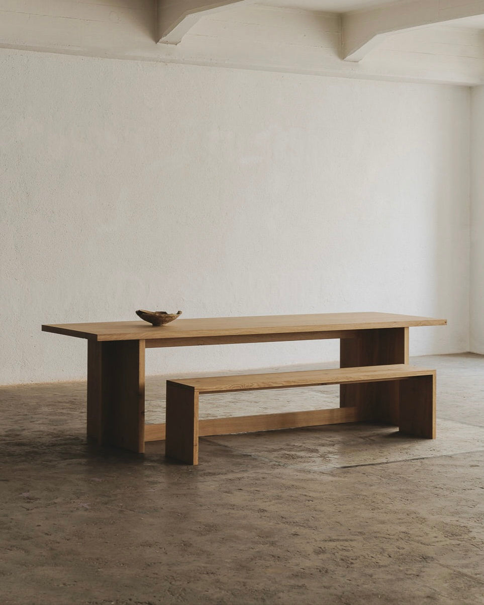 A minimalist, spacious room showcases the Paris Dining Table from ASERIES OF OBSTACLES, SL with a matching bench for social gatherings. A small decorative bowl accents the table against textured concrete floors and white walls, with an exposed beam. Natural light enhances French elegance.