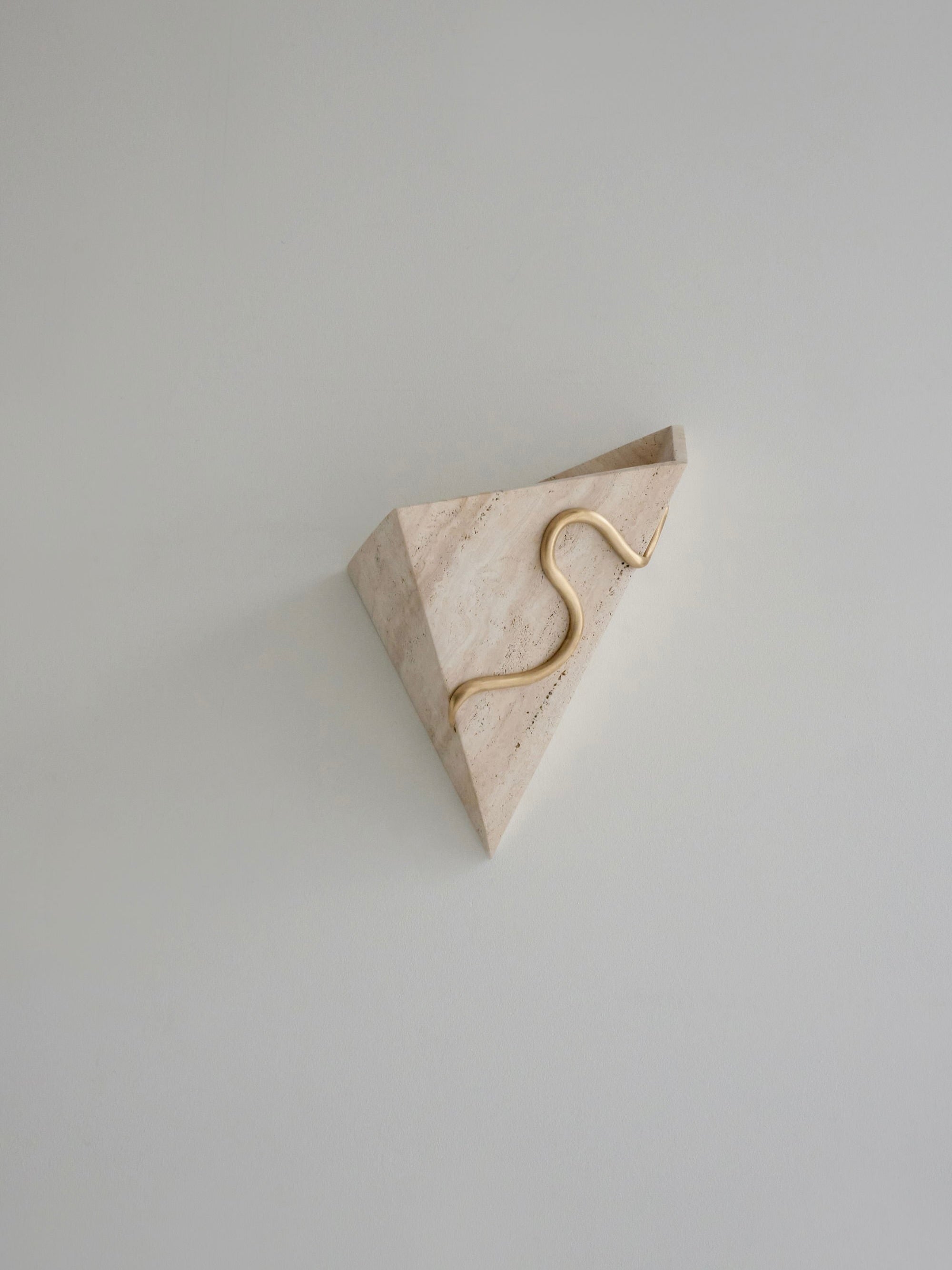 The Therapeutic Wall Light 1 by Mariza Galani is a wall-mounted, triangular stone light fixture that features a sleek, wavy metallic accent running diagonally across its surface. This marble wall light contrasts the natural stone texture with the polished metal, creating an artistic and modern look against a plain background.