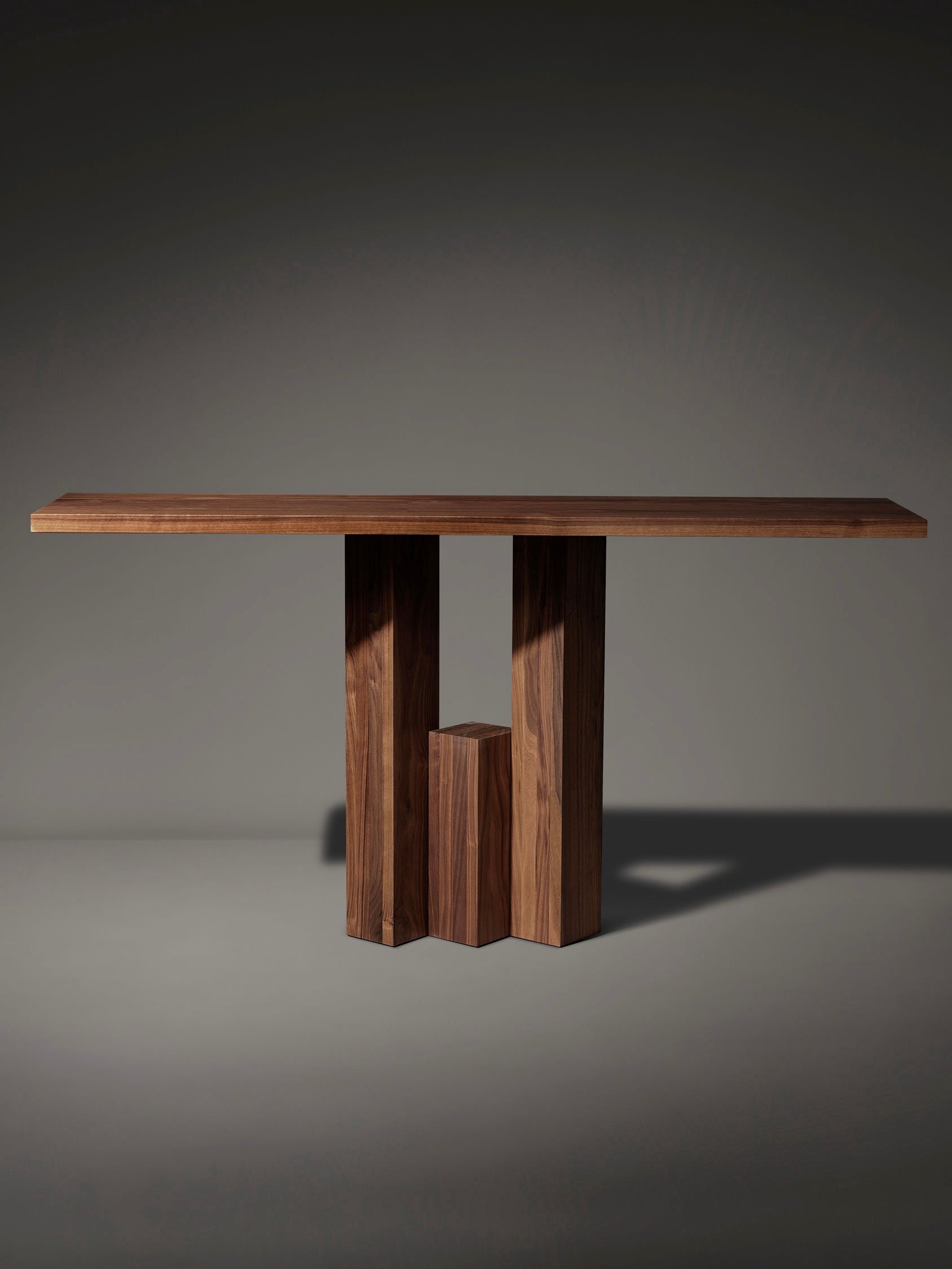 Introducing the Mokko Fenestra Console, a contemporary wooden table featuring a flat top and an abstract geometric base. Its three vertical supports of varying heights create artistic shadows on a gray minimalist background, offering a customizable design ideal for any space.