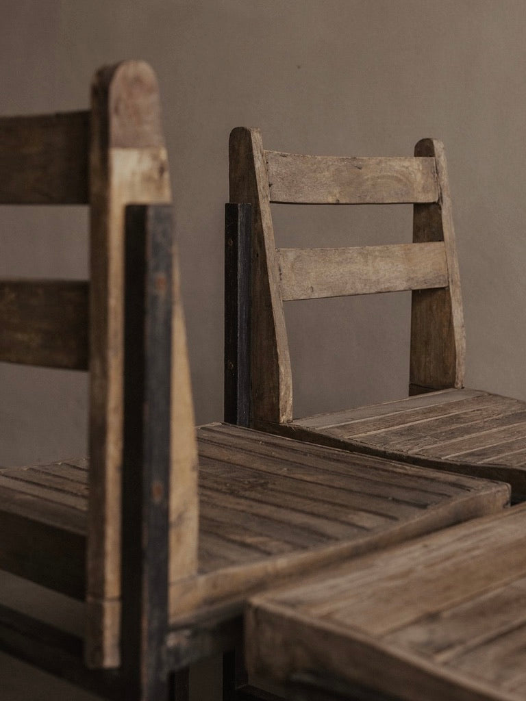 Two rustic chairs by Bicci de' Medici stand before a taupe wall. Resembling the authentic PJ-SI-65-A Pierre Jeanneret chair, they feature simple, straight backs and teak slat seats, exuding a vintage, minimalist charm.