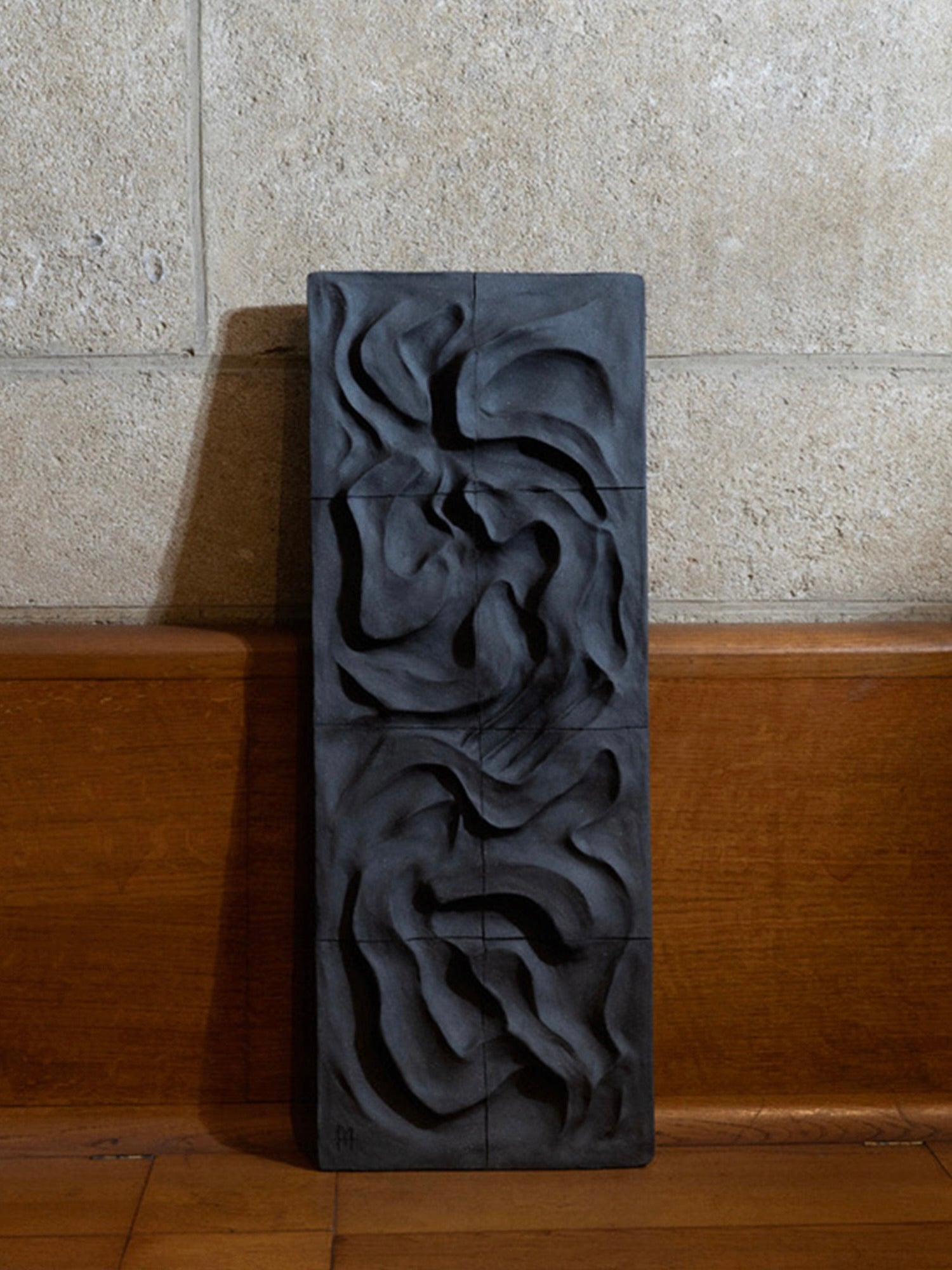 The Kilauea Lava rectangular ceramic wall sculpture by Franck Scala leans against a wooden bench and stone wall. Its textured black surface with swirling, carved patterns adds depth and shadow to enhance its abstract design.