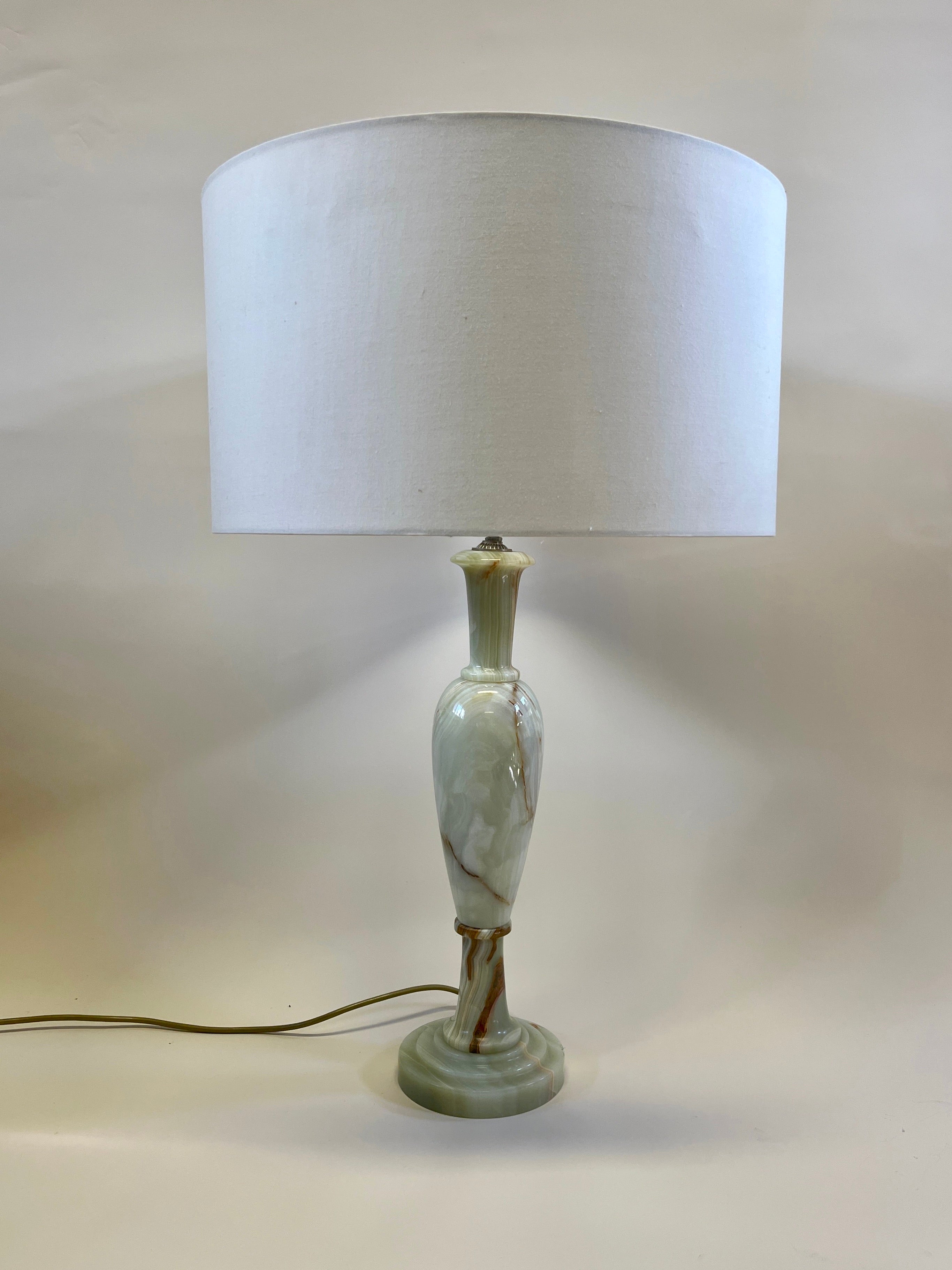 The Large Vintage Onyx Lamp by Maud Vaughan showcases an elegant onyx base with a white cylindrical lampshade. The lamp's design features a slender, decorative onyx stem with subtle green and brown veining, leading to a circular base. The lamp is illuminated, casting a soft glow from the antique gold flex and rewired brass fitting.