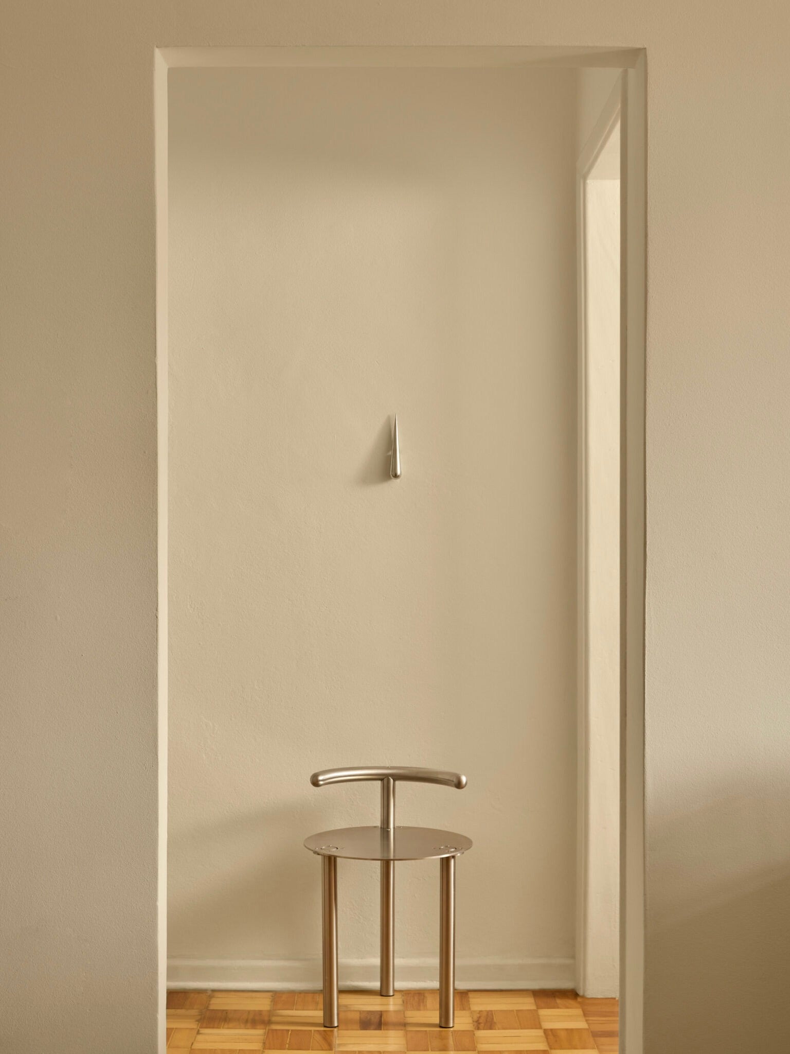 A minimalist room features a sleek, metallic chair crafted from cast aluminum with a curved backrest sitting on a wooden floor. Centered above the chair against a plain, cream-colored wall is the "Gota" Bottle Opener by Pedro Avila, adding a small, droplet-shaped design accent to the decor. Natural light softly illuminates the scene.