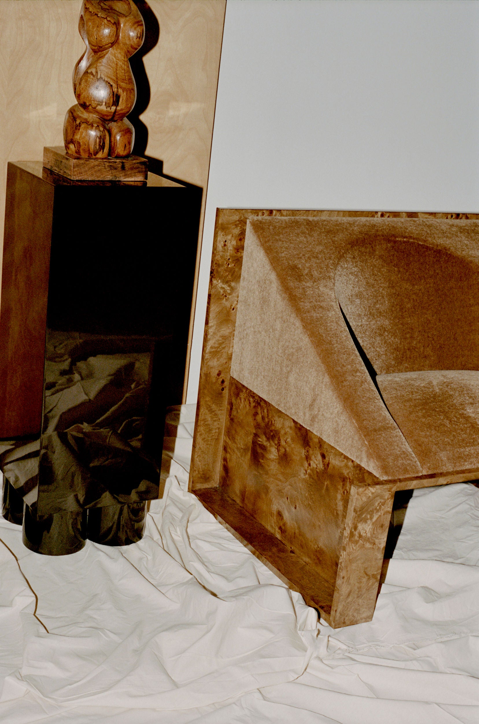 A modern scene presents a wooden sculpture on a black pedestal next to the 'Perceptions' Armchair by Known Work, featuring geometric wood and velvet upholstery. The floor is adorned with a crumpled white sheet against a minimalist neutral backdrop.