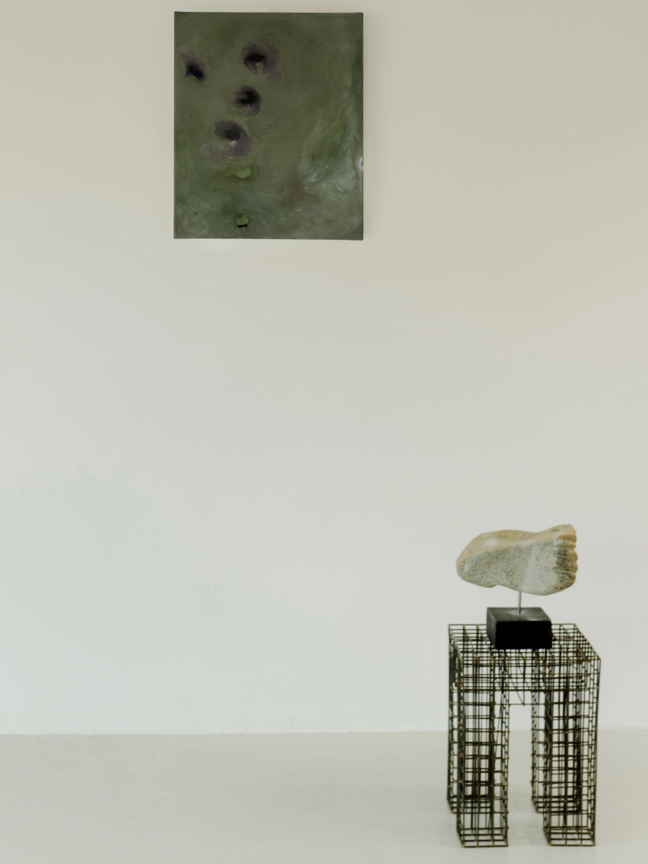 A minimalist art gallery displays "Let go of gravity" by Yeliza Gevorgyan, a painting with abstract green hues using natural pigments on a white wall. Below, a stone face sculpture sits on a black stand atop a wireframe pedestal.