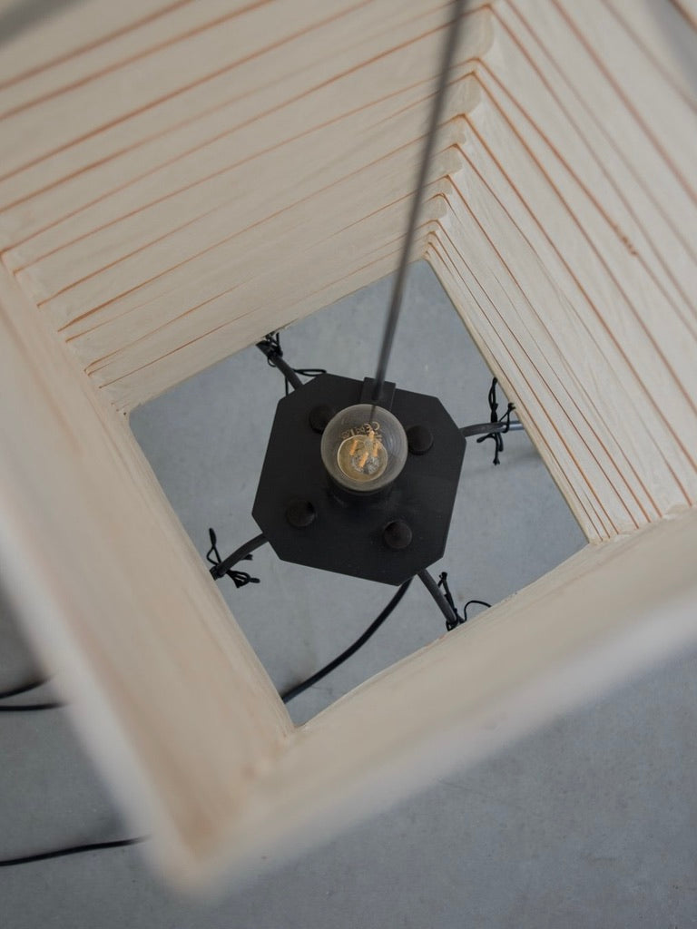 Viewed from above, this square wooden lamp with vertical slats evokes a mid-century modern aesthetic. Encased in a black metal framework, its round bulb warmly glows against the smooth gray surface below, reminiscent of Bicci de' Medici's Isamu Noguchi - Akari UF vintage lamp.