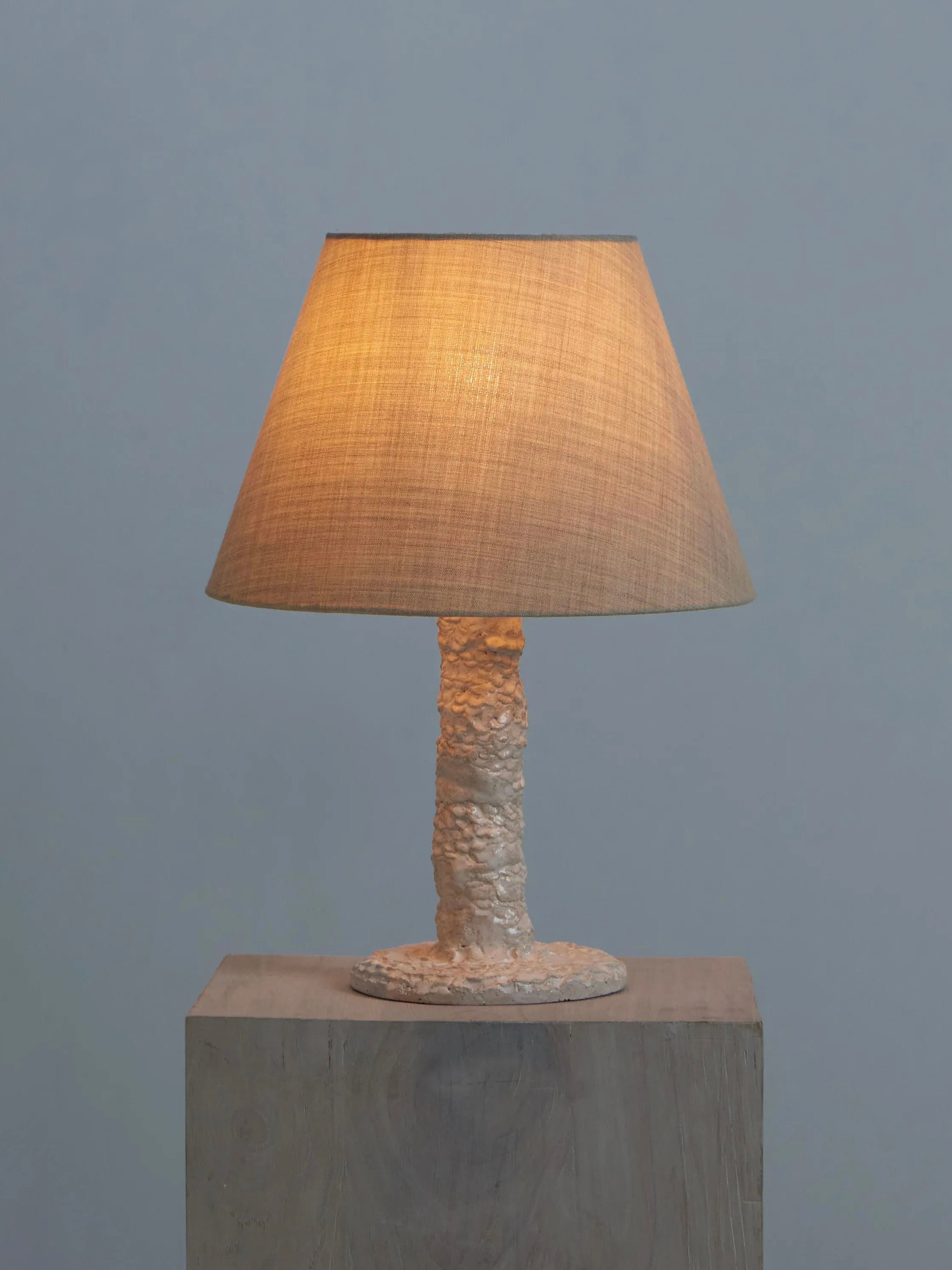 The Casa Bedside Light by Project 213A is a handmade table lamp with a beige textured base and conical woven lampshade, emitting a warm glow. It rests on wood against a soft blue-gray background, showcasing bespoke dimensions for an artistic touch.