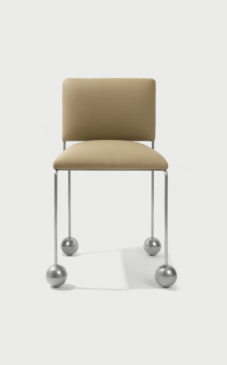 The 'Ball Foot' Chair by Panorammma boasts a beige vegan textile cushioned seat and backrest. It elegantly stands on thin metal legs with unique large round metallic feet, set against a plain light gray background that complements its modern and sophisticated design.