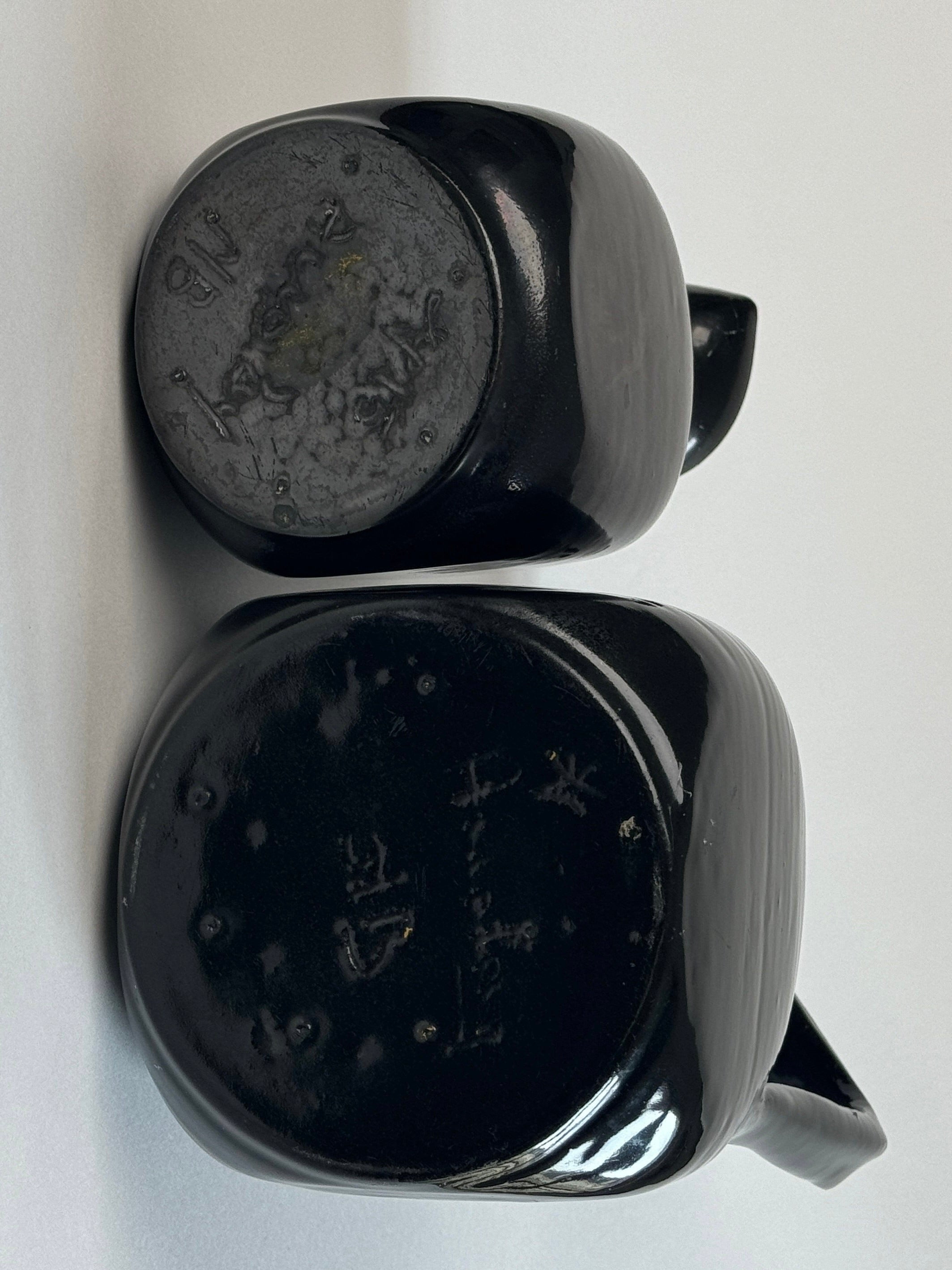 Two black glazed carafes with handles, from Figuresdesign's Pair of Accolay Ceramic Carafes collection, are placed upside down, displaying their bases. The base of each carafe shows engraved or embossed markings or characters, though they are difficult to read due to the lighting and angle. The background is a plain surface.