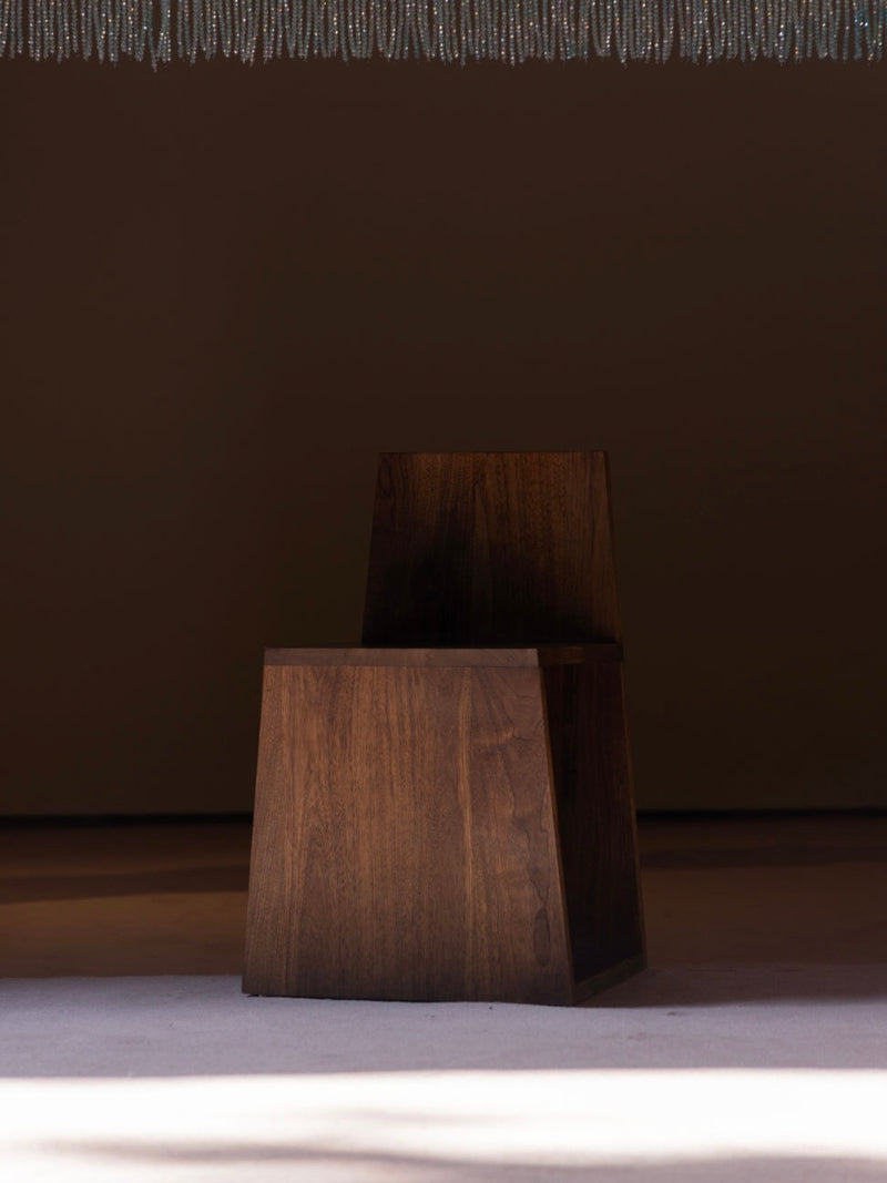 A 'Totem' Chair by Siete Studio, made from solid walnut, is positioned on a carpeted floor. Its geometric design and clean lines are enhanced by the dim lighting. A beaded curtain subtly appears above, adding a totemic element to the scene.