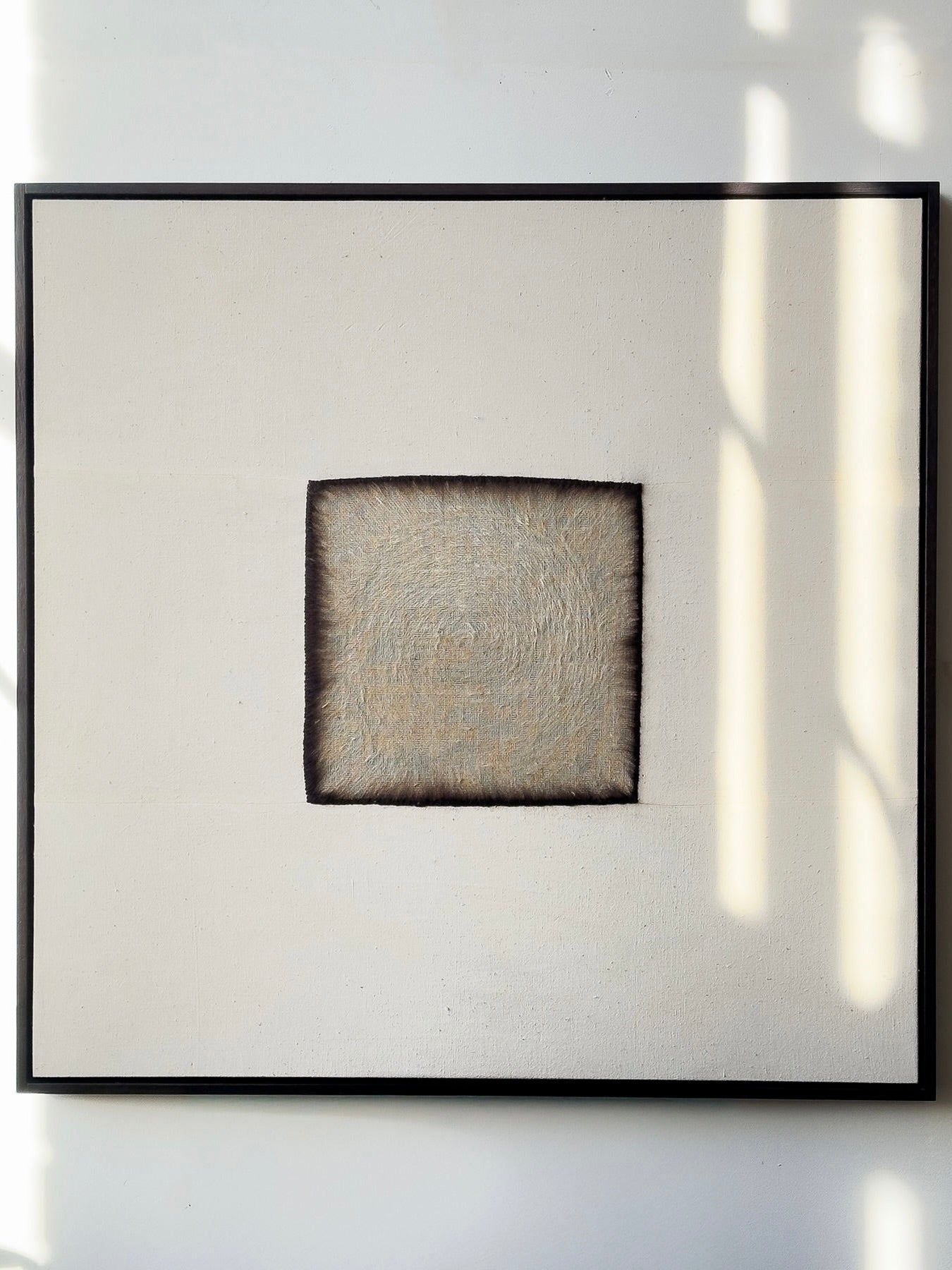 Charred," a square painting by Bec Kirby, features a textured fuzzy central square with darker blurred edges and a lighter interior, surrounded by a plain off-white background. It is framed in black solid oak, with soft shadows falling from the right.