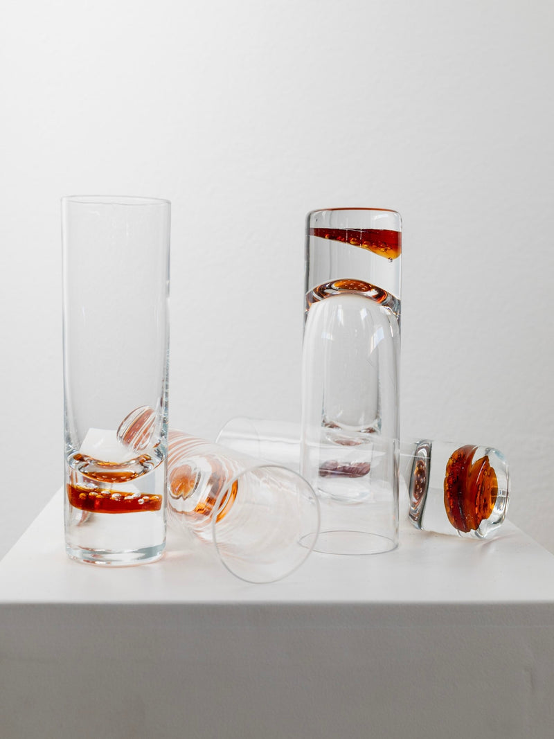 The arrangement of Bottega Jacobs' 5 Retro Murano Drinking Glasses, featuring tall, transparent designs with round, amber-colored bases, creates a vintage charm on a white surface against a plain backdrop. Some glasses stand upright while others rest on their sides, evoking memories of classic Murano aesthetics.