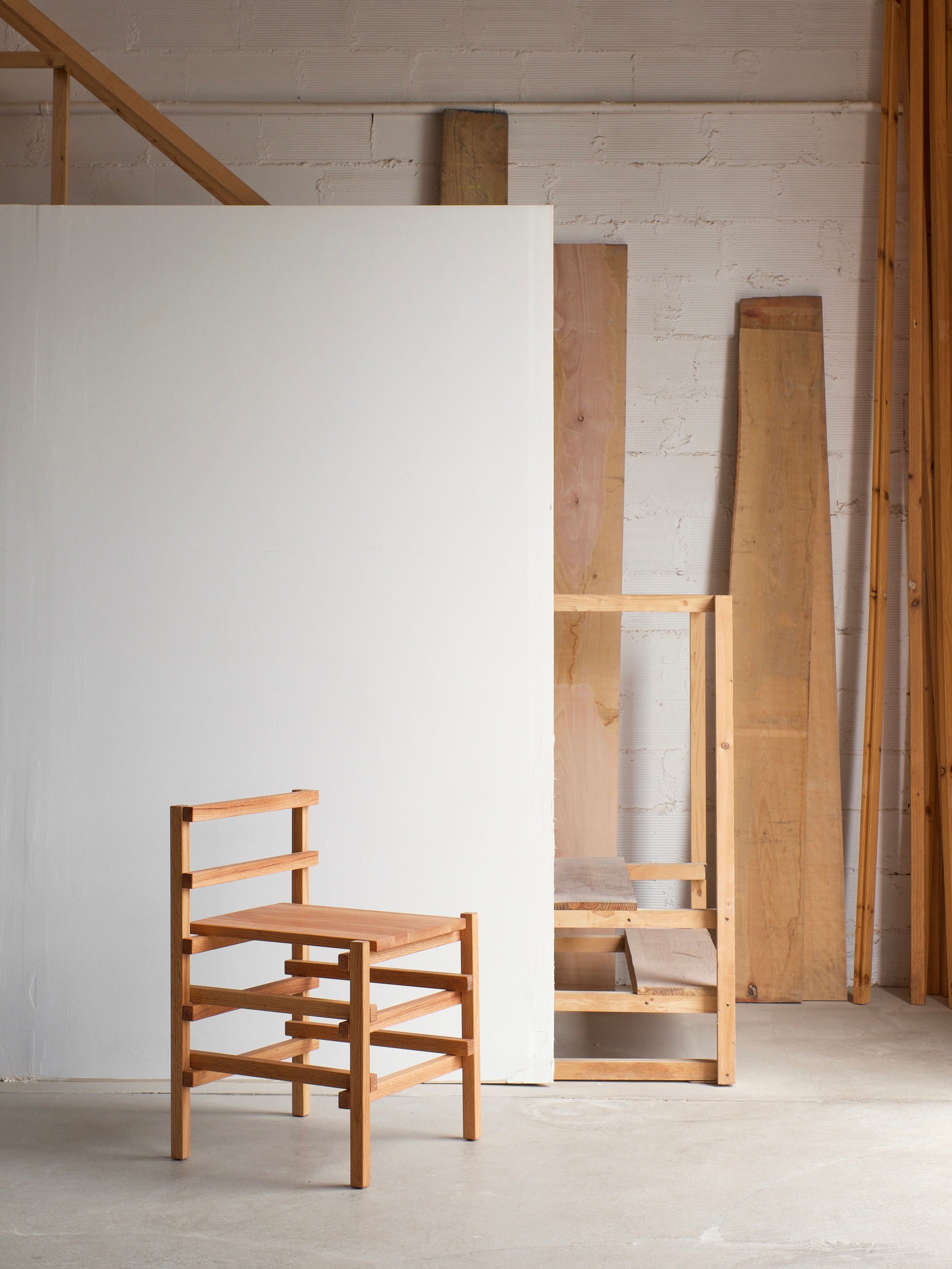 A Small Palma Chair by malva, exuding modern elegance, stands on a concrete floor in front of a white-paneled wall within a workshop-like space. Various wooden planks and beams lean against the wall in the background, contributing to the rustic, industrial ambiance while underscoring its versatile statement piece quality.