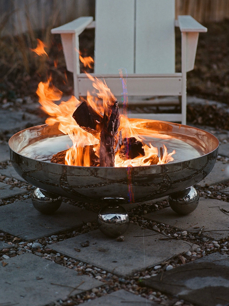 A cozy scene featuring the MUHLY Flecto Fire Bowl, a polished stainless steel fire pit with burning logs set ablaze with bright orange flames on a patio. In the background, there's a white Adirondack chair. The surrounding area is enhanced by paving stones and scattered pebbles, completing the elegant design.
