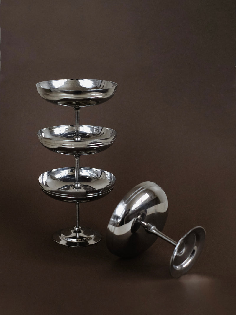 A set of four Ice Coupes by septembre studios stands elegantly, each reflecting light against a dark brown background. Crafted in France, one coupe rests on its side, highlighting its smooth surface—ideal for cradling a scoop of ice cream.