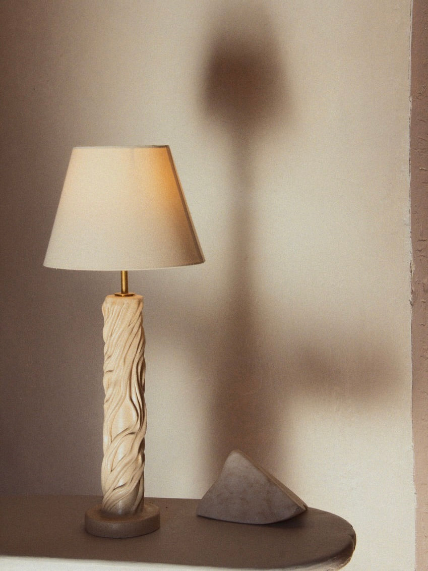 The "Waves" Table Lamp by Lorea Palacios features a ceramic base with intricate carvings and a white shade. It stands on a light surface next to a small rock, with its matte glaze adding charm as it casts a soft shadow on the beige wall—ideal for minimalist interiors.