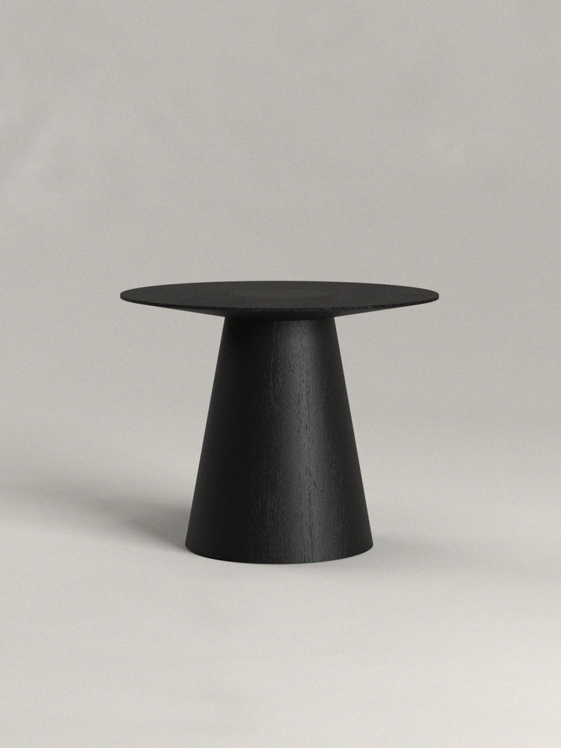 The Totem Side Table by Siete Studio is a round black piece with a conical base, showcased against a neutral background. Its minimalist design features clean lines and a matte finish, crafted from natural materials that offer elegance and simplicity.