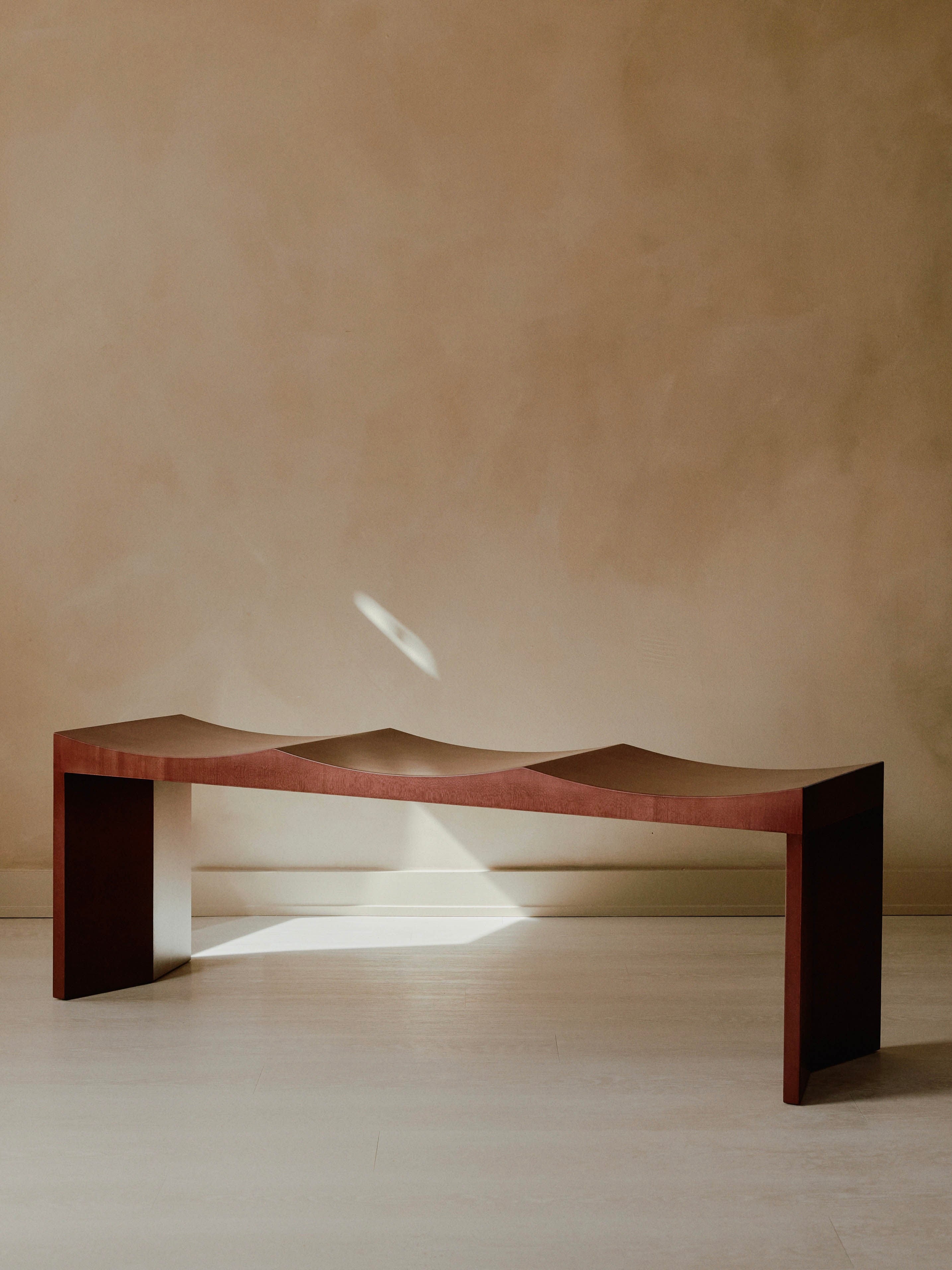 A minimalist Arc Bench from Juntos Projects, featuring chevron legs and a wavy seat design, is placed in front of a beige wall. Sunlight casts a small bright spot on the wall and part of the bench, creating a serene and contemplative atmosphere.