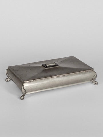 The Art Deco Metal Box by Spigel is a rectangular piece showcasing vintage Scandinavian craftsmanship. It features a hinged lid and small, curved feet at each corner. Resting against a plain background, its slightly worn appearance is complemented by a simple handle on top, evoking the elegance of polished pewter.