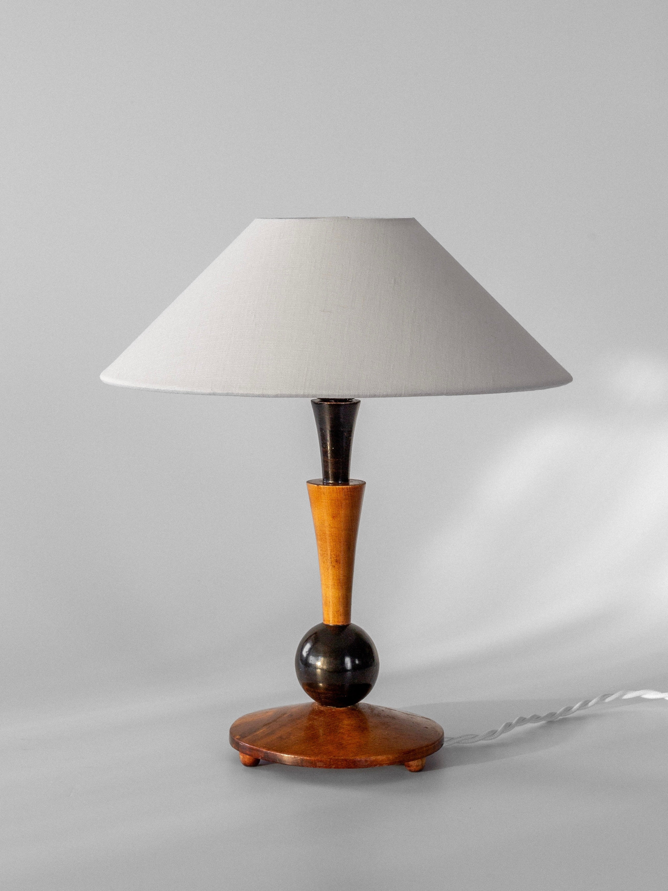 The Art Deco Wooden Table Lamp from Spigel offers a stylish vintage character with its wide white lampshade and a wooden base, complete with a spherical design near the top. Echoing the essence of 1935 Sweden, this lamp is set against a plain gray background, featuring a white cord extending from the base.