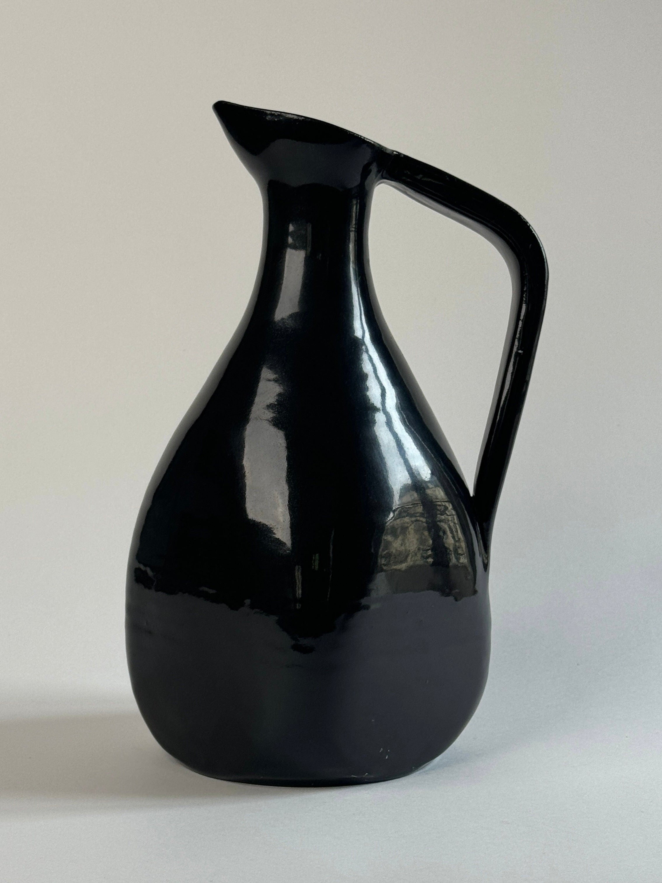 On a plain white background, a minimalist black glazed ceramic carafe from the Figuresdesign brand catches the eye with its glossy finish. The curved body, narrow spout, and handle extending from top to middle are reminiscent of vintage French pottery, evoking the charm of Accolay France pitchers. The glossy surface reflects light beautifully, creating highlights on this elegant piece.