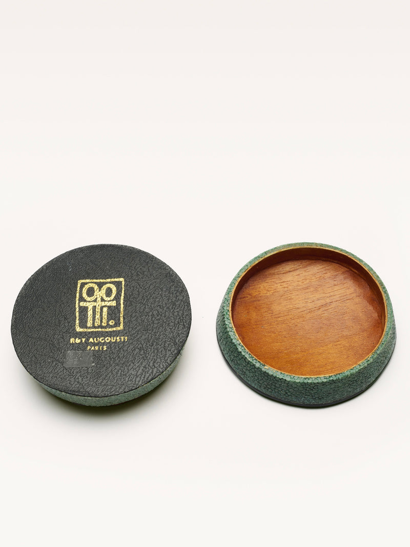 A round black Small Shagreen Pearl Box by Galerie MLS, with gold logo, sits open beside a shallow brown wooden dish with a green textured rim, set against a plain light-colored background.